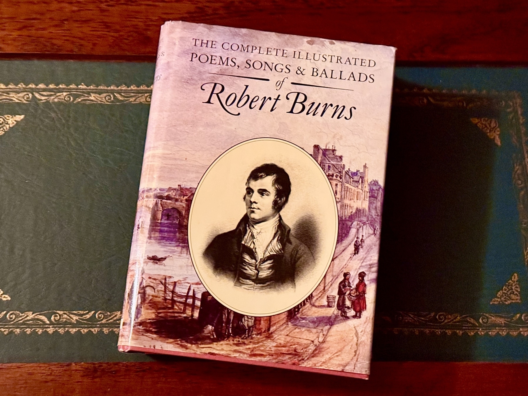 A book titled "The Complete Illustrated Poems, Songs & Ballads of Robert Burns" is placed on a decorative surface. The cover features an illustration of Robert Burns in an oval frame, set against a background depicting a historic scene with buildings and people.