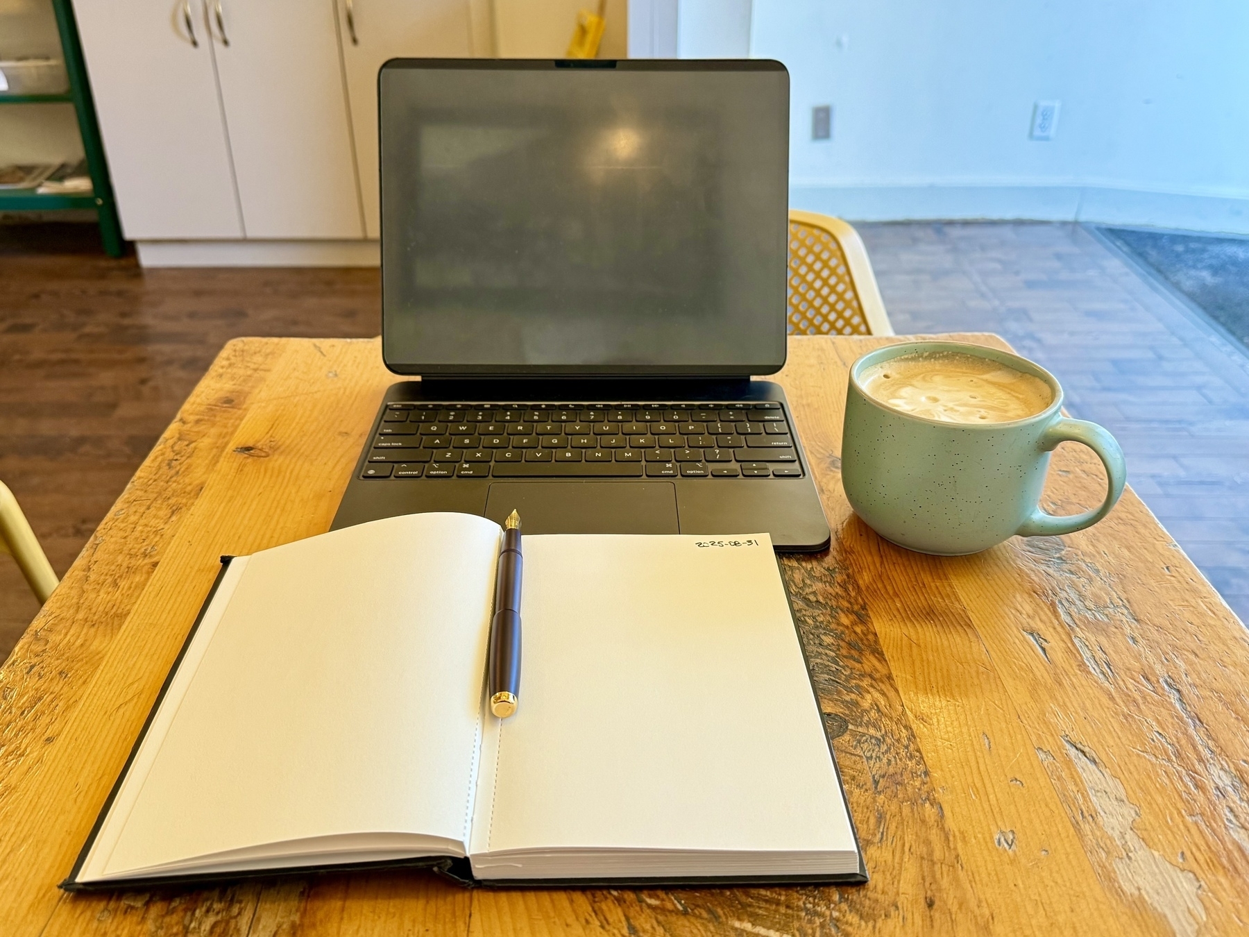 A laptop sits on a wooden table with a blank notebook and pen beside it. A large mug filled with a frothy drink is also on the table. The setting appears to be a cozy room with wooden flooring, white cabinets in the background, and a yellow chair partially visible.