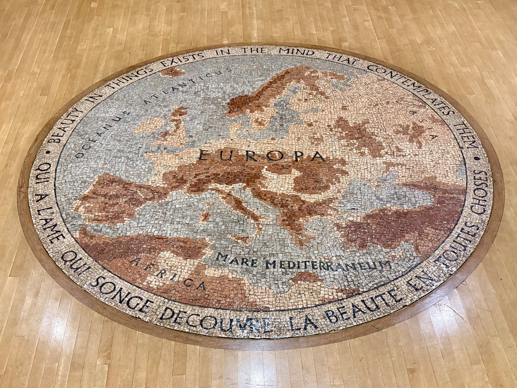 A circular mosaic floor piece featuring a map of Europe with adjacent regions of Africa and the Mediterranean Sea. The mosaic employs earthy tones with shades of brown, beige, and gray. Surrounding the map is an inscription that reads: "Beauty in things exists in the mind that contemplates them. A l'âme qui songe découvre la beauté en toutes choses." The artwork is set on a wooden floor.