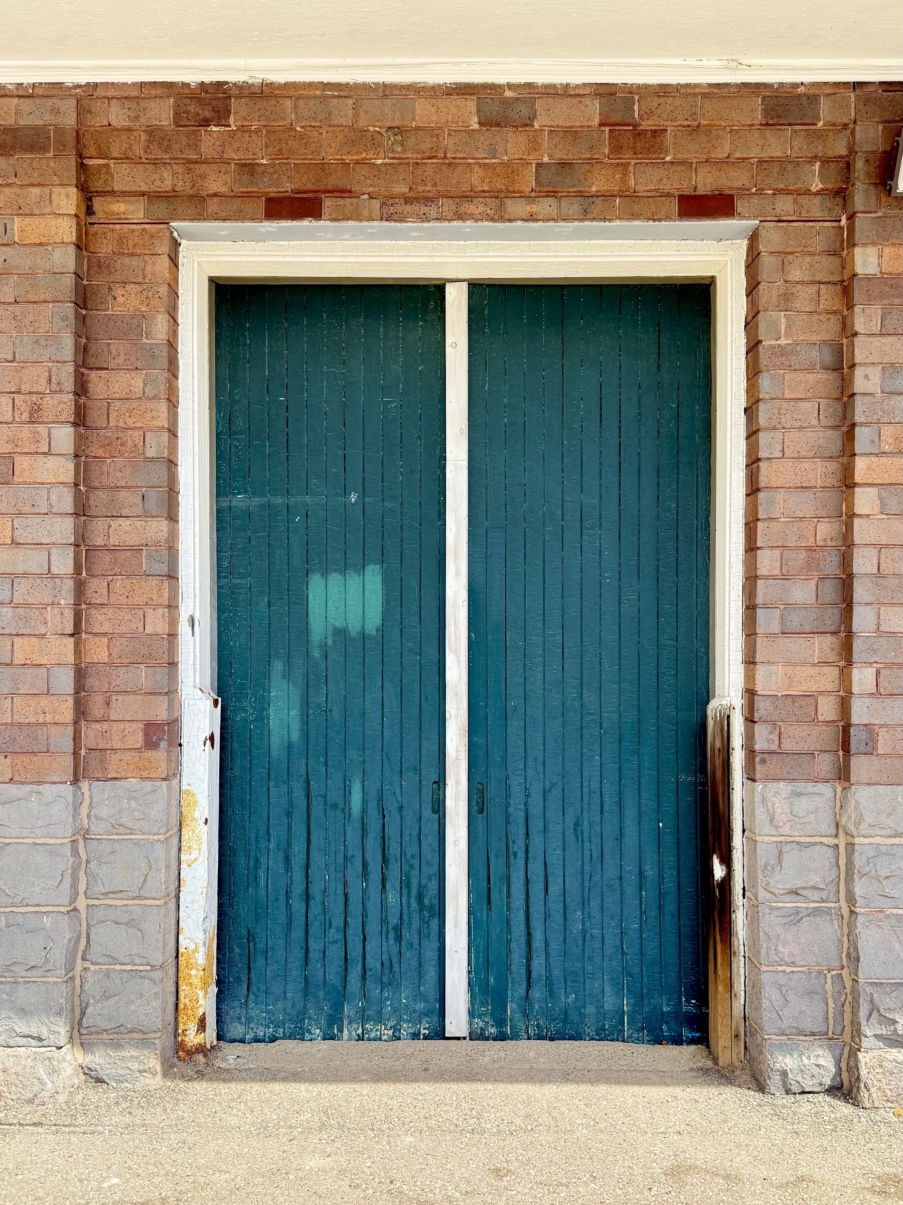A pair of tall, blue wooden doors are set within a brick wall. The surrounding bricks are reddish-brown, with a light stone base. The doors show signs of age, with some peeling paint and wear. The scene suggests an entrance to an old building.