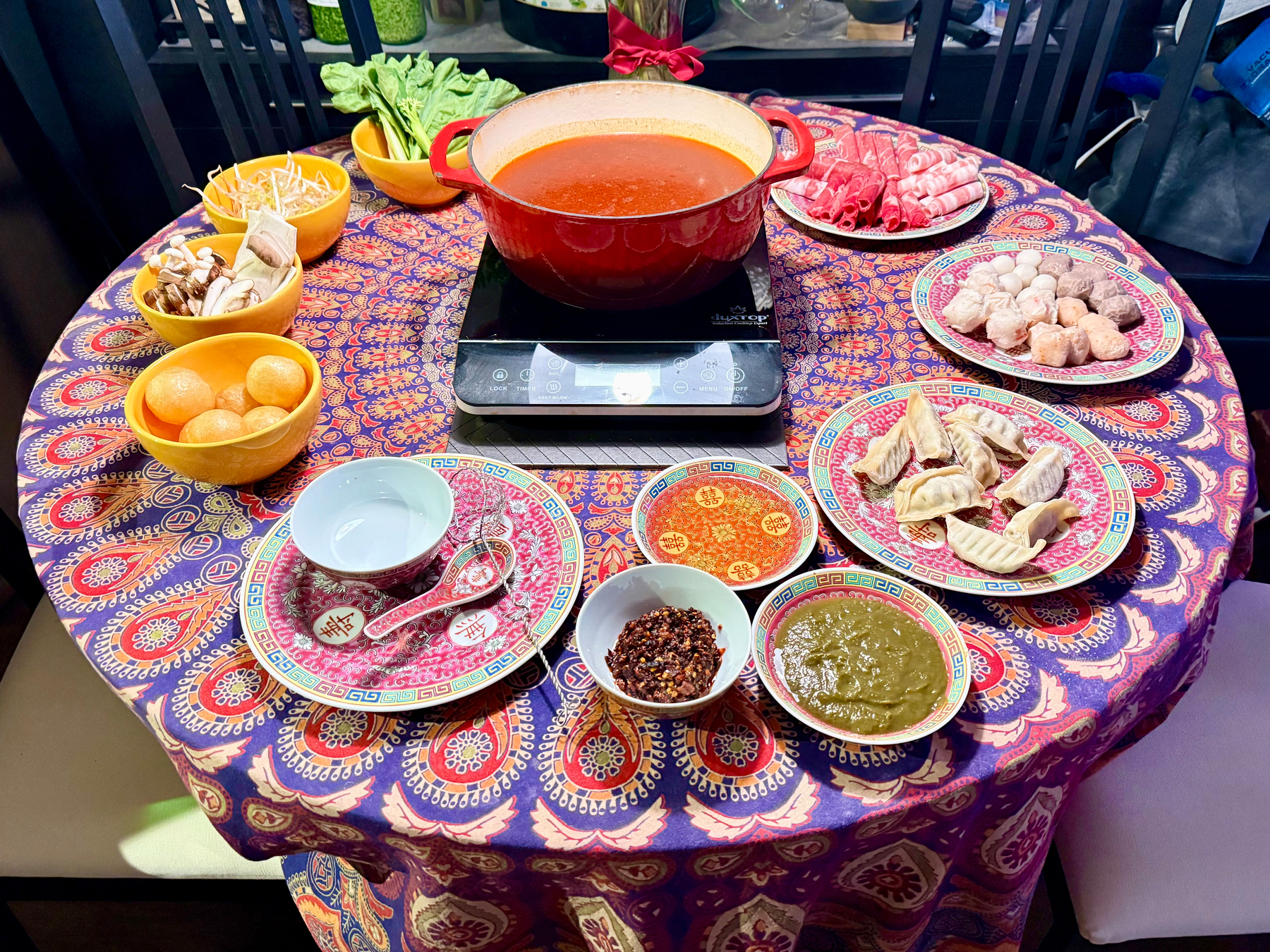 A vibrant table setting features a pot of simmering broth surrounded by various ingredients and condiments for hot pot, including sliced meats, dumplings, vegetables, sauces, and gluten balls, all on a decorative tablecloth.
