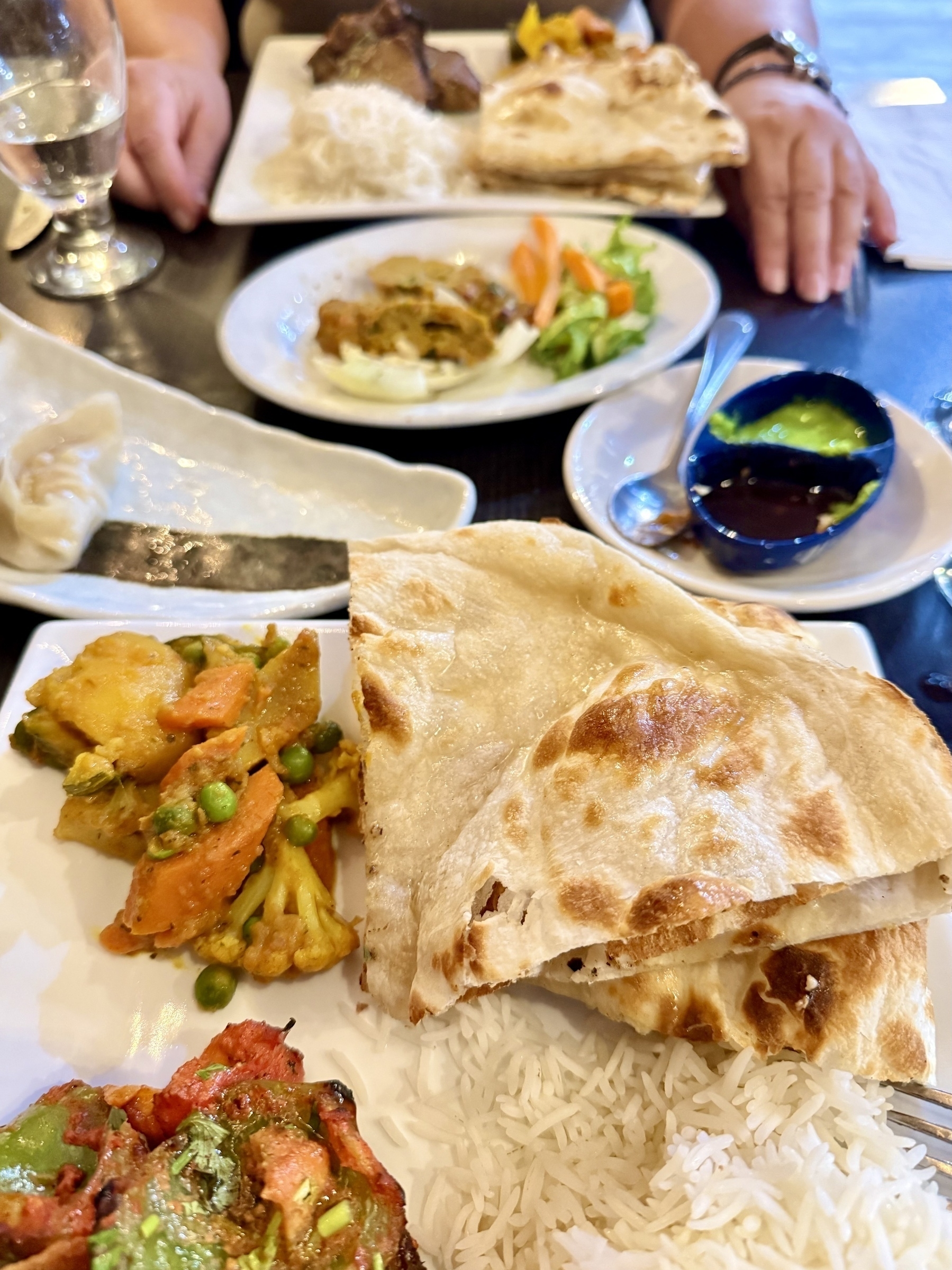A close-up of a plate featuring Nepalese cuisine. The plate includes fluffy basmati rice, pieces of naan bread, a vegetable curry with carrots, peas, potatoes, and cauliflower, and a vibrant vegetable dish. In the background, there's another plate with more naan, a piece of meat, and salad. A small dish contains green and dark brown sauces with a spoon. A glass of water and a person's hands are also visible on the table.