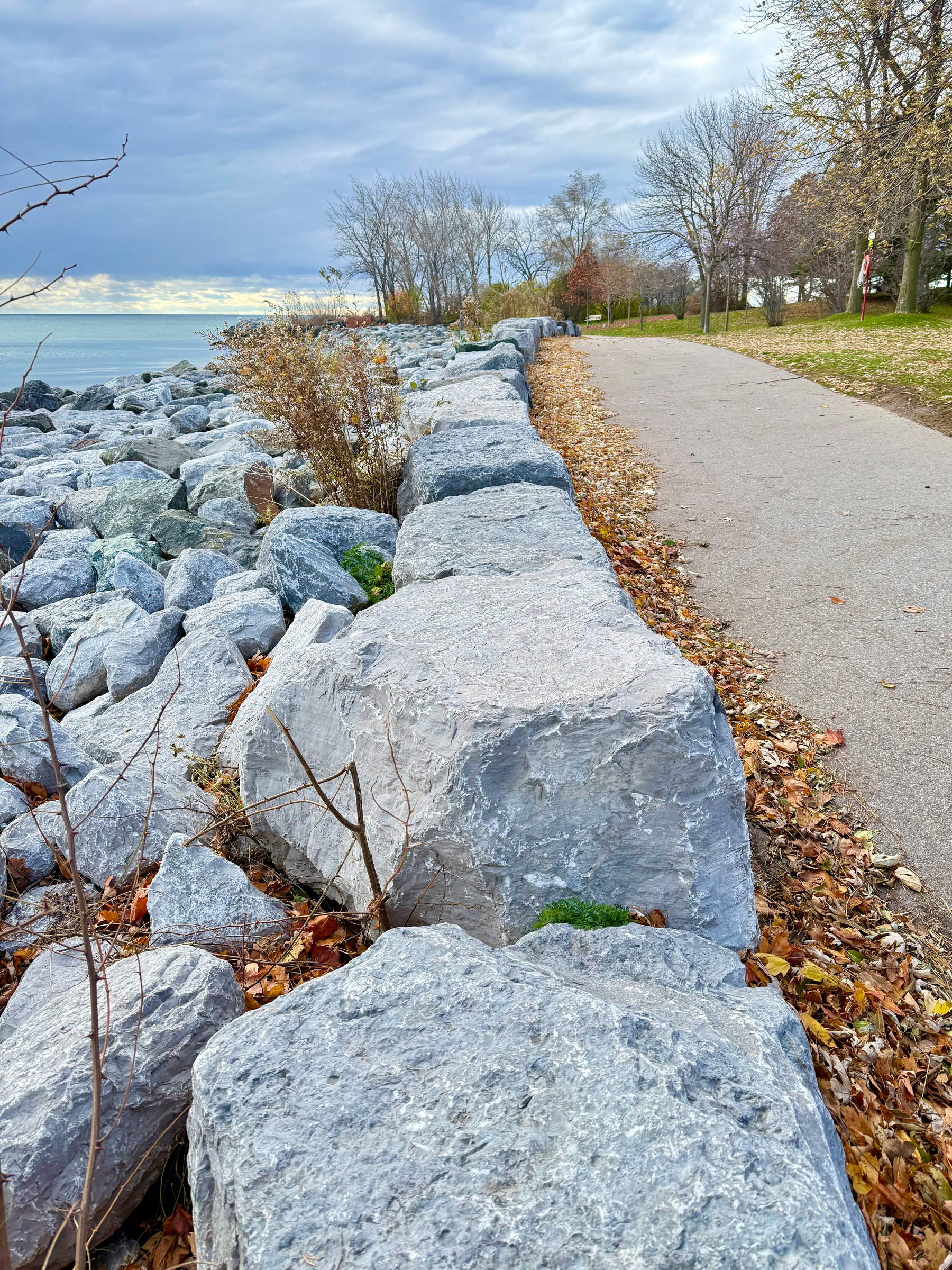 A scenic view of a lakeside path bordered by large gray rocks. The path is lined with fallen leaves, and bare trees can be seen on the right. To the left, the rocks continue toward a calm body of water under a cloudy sky. The colors suggest an autumn setting.