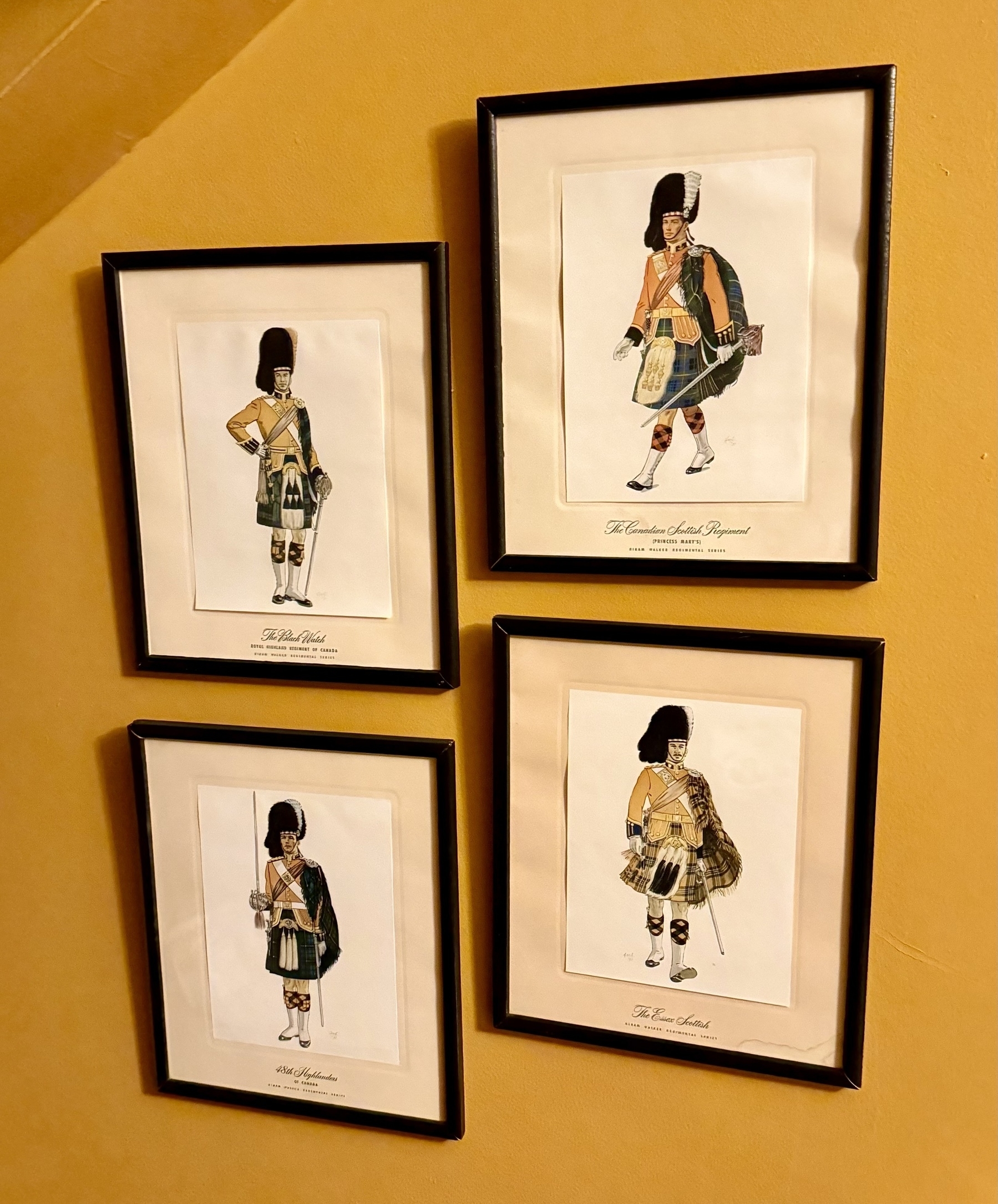 Four framed illustrations hang on a yellow wall, each depicting soldiers in traditional Scottish military attire. The images feature distinctive elements such as kilts, sporrans, and hats. The frames are simple with black borders, and each illustration has a caption beneath, identifying the specific regiment or type of attire displayed.
