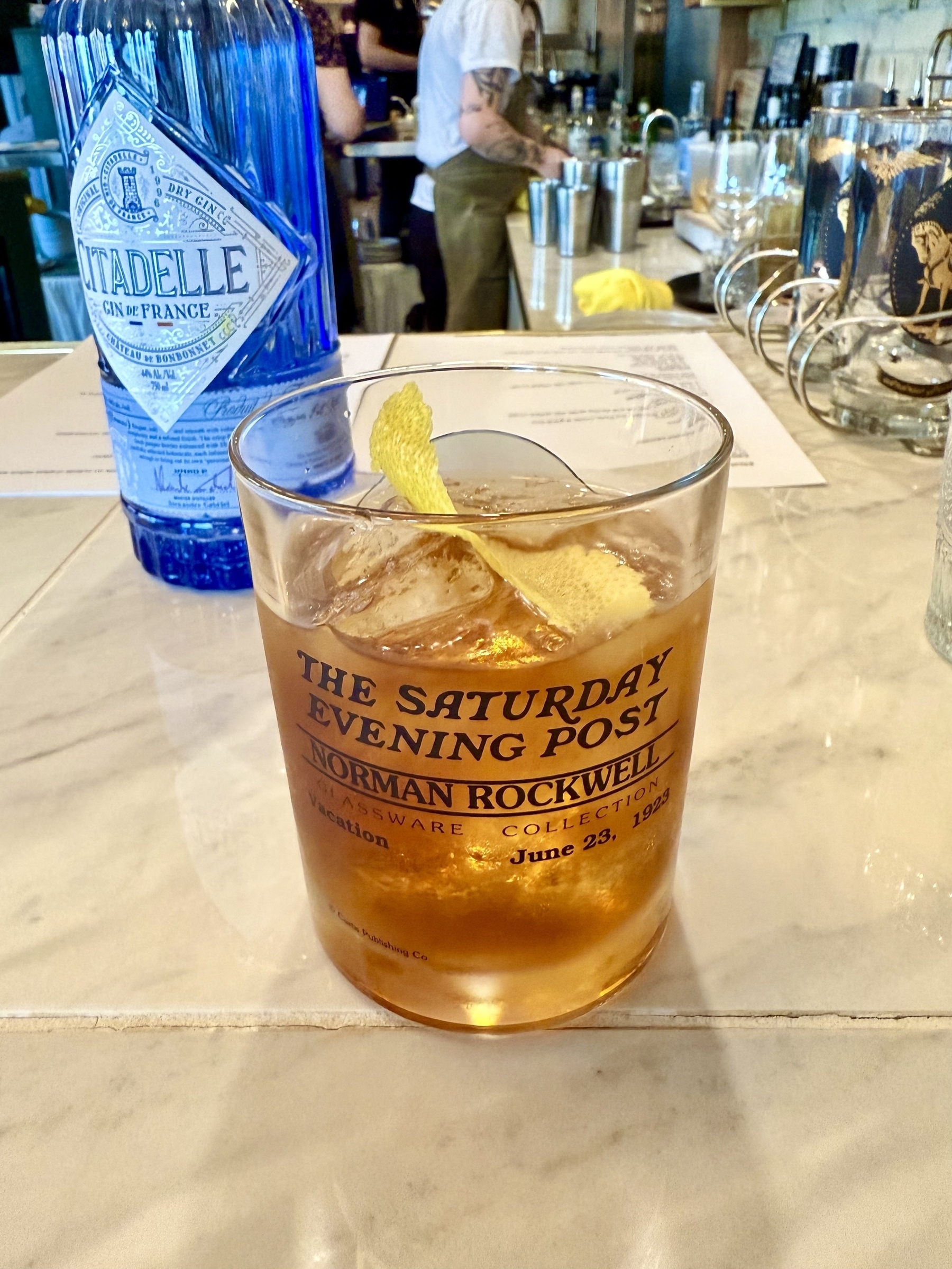 A glass filled with an amber-colored cocktail garnished with a lemon twist sits on a marble bar counter. The glass has text referencing "The Saturday Evening Post" and "Norman Rockwell." Behind it, there is a blue bottle labeled "Citadelle Gin de France." In the background, a person in a white shirt and apron is working at a bar with various bottles and utensils visible.