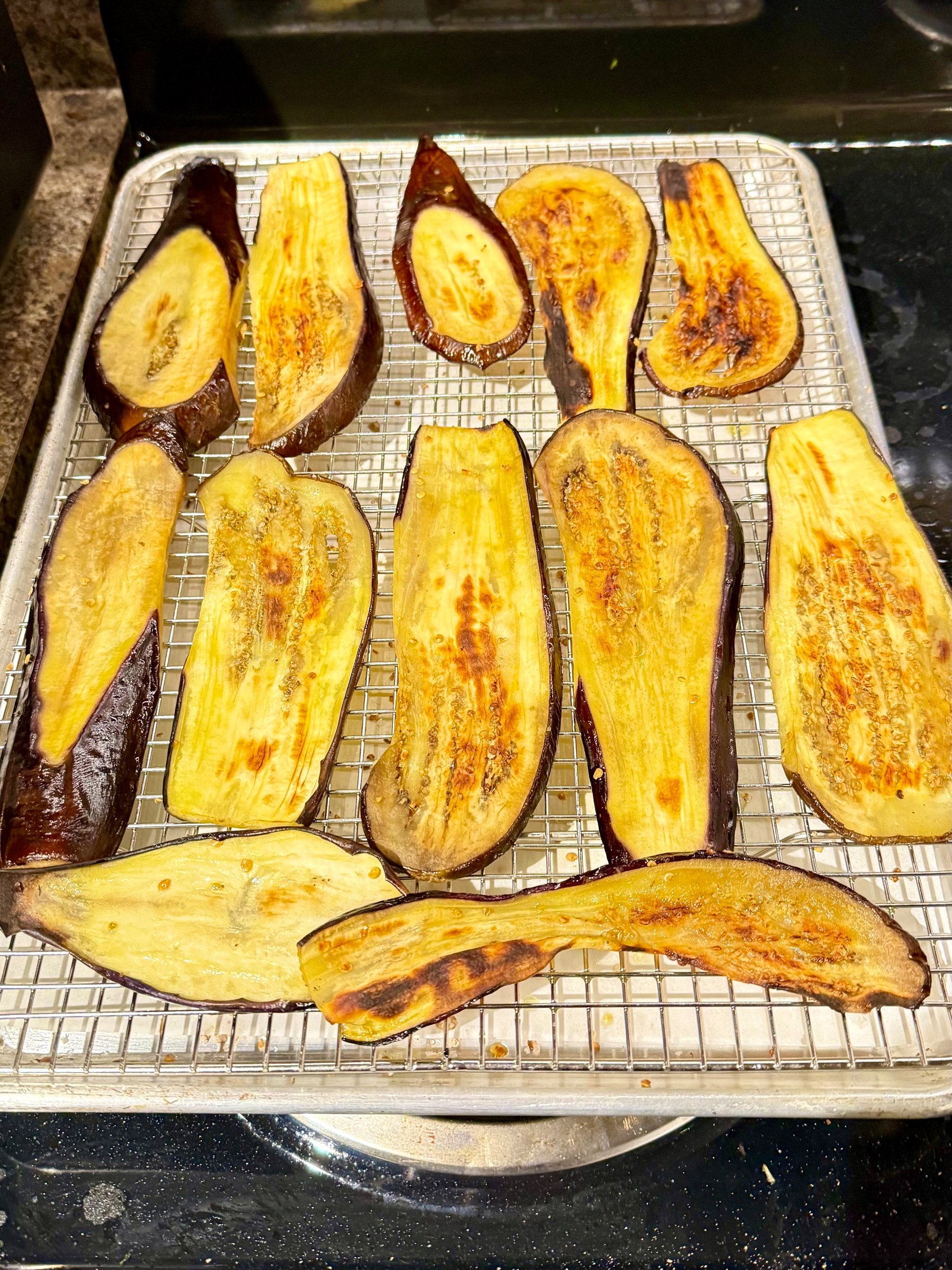 Roasted eggplant slices arranged on a metal rack over a baking sheet. The slices are golden brown with darkened edges, indicating they are cooked. The rack is set on a dark countertop, contrasting with the light colors of the eggplants.
