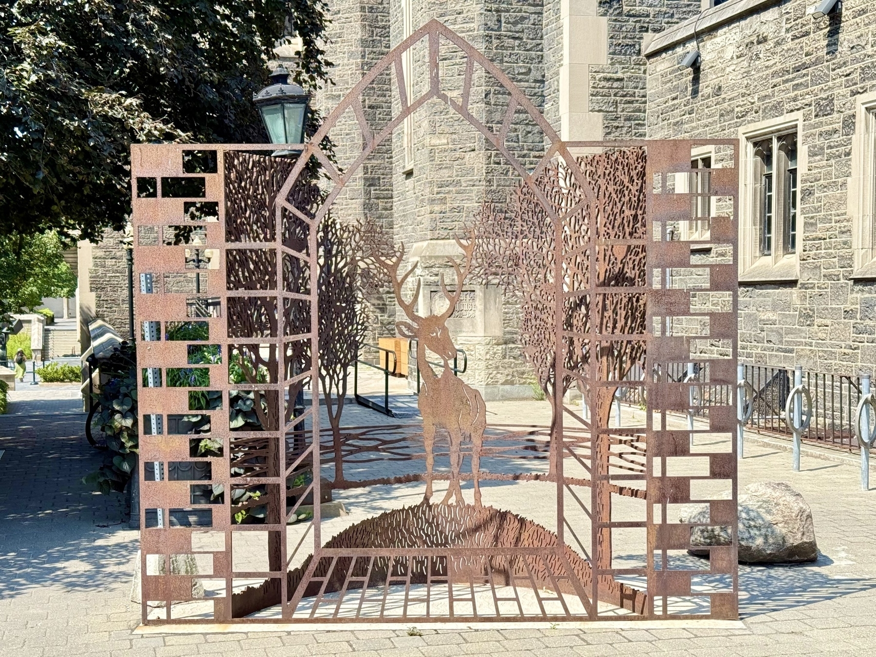 A metal sculpture of a stylized deer stands in the center of a decorative archway made from rust-colored metal. The design includes cut-out patterns resembling trees and branches. In the background, there is a stone building and surrounding greenery, with sunlight casting shadows on the pavement. The scene is peaceful and artistic, integrating nature and architecture.
