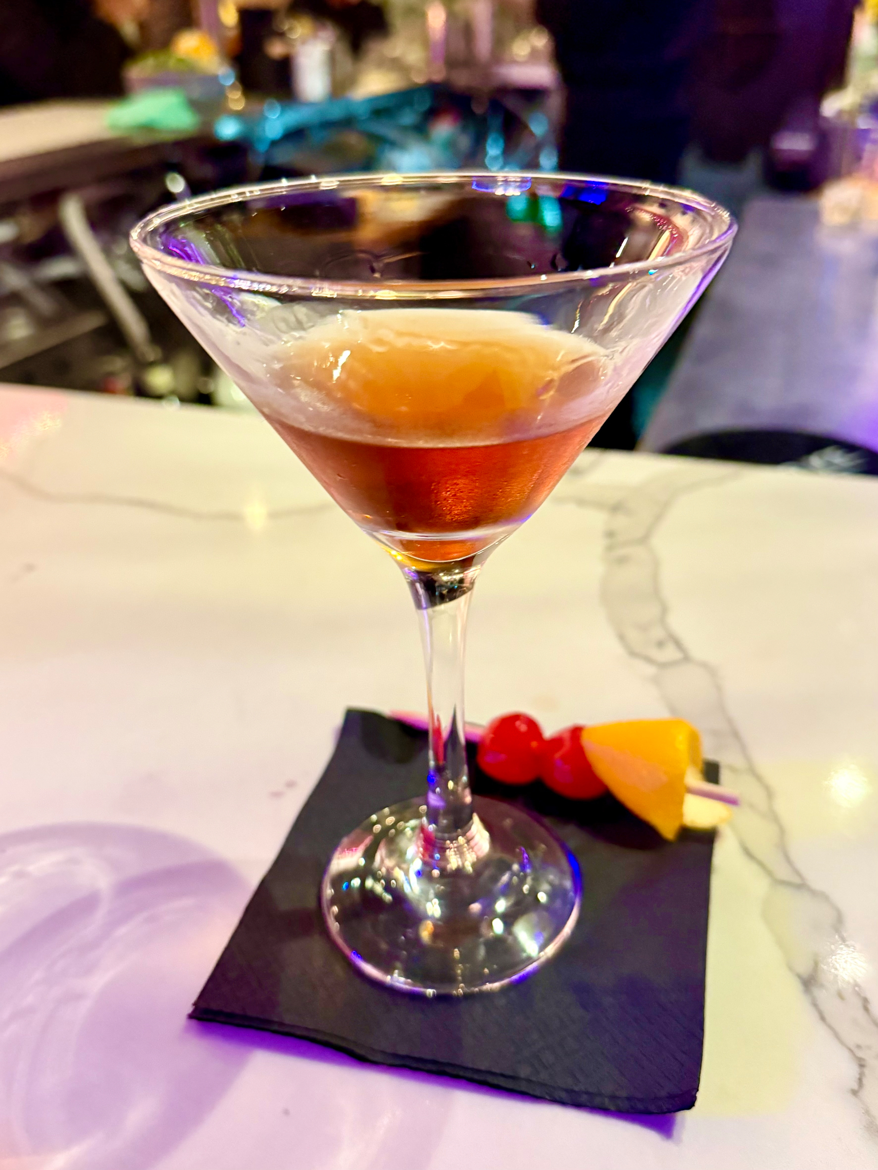 A martini glass with an amber-colored cocktail on a black napkin on a marble table. Next to the glass is a fruit skewer with two red cherries and a slice of orange. The background is softly blurred, showing a dimly lit bar environment.