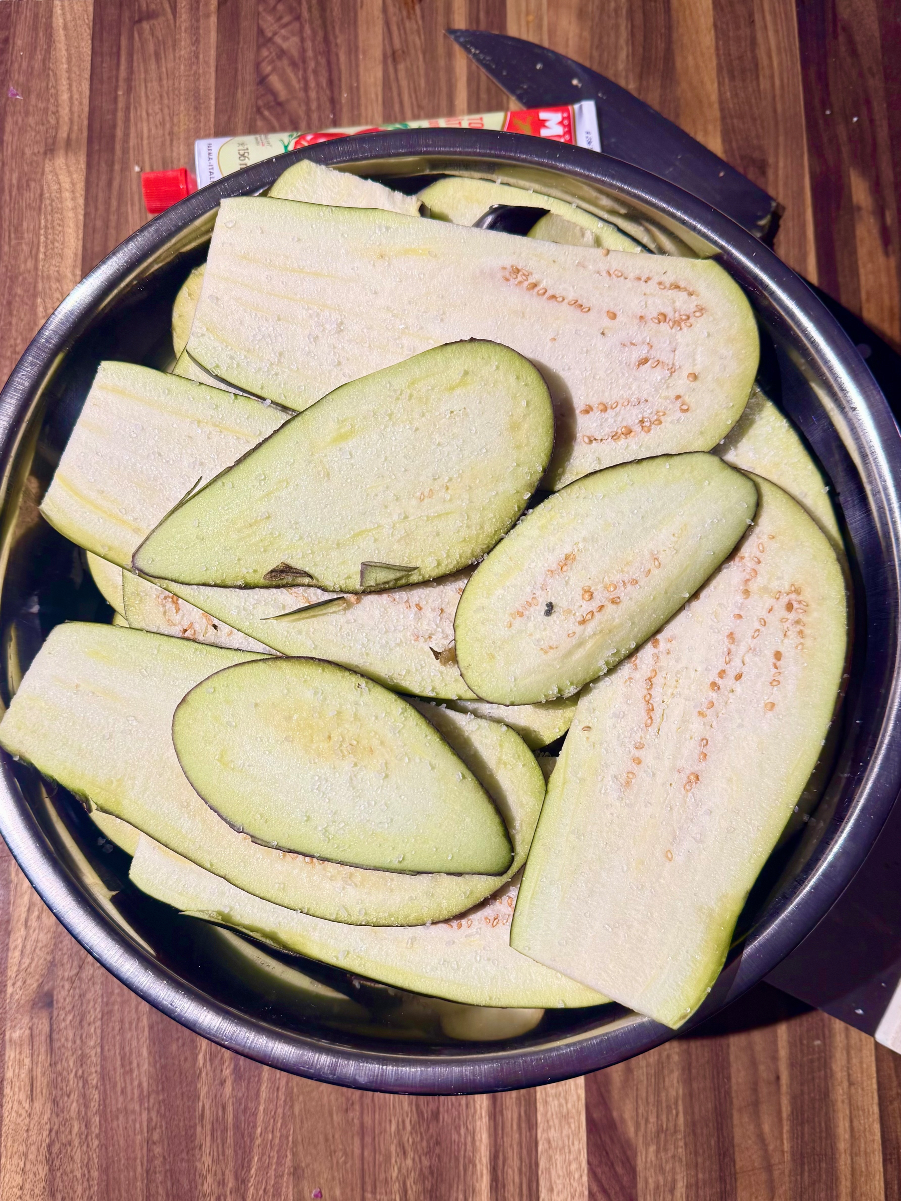 A metal bowl filled with thinly sliced eggplant, lightly salted. The slices are pale green with visible seeds. In the background, there's a partially visible knife and a tube of tomato paste on a wooden surface.