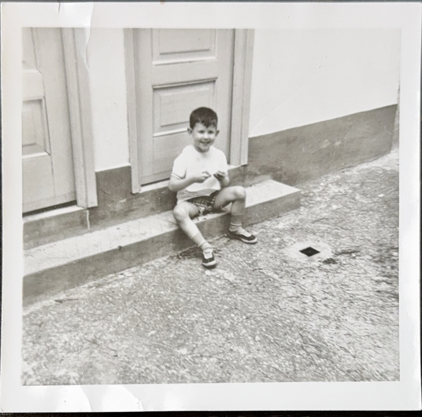 A black and white photo of a young boy sitting on a doorstep. He is wearing a light-colored shirt, shorts, and shoes with socks. The boy is smiling and holding something in his hands. The setting appears to be outside a building with two doors visible. The ground is paved.