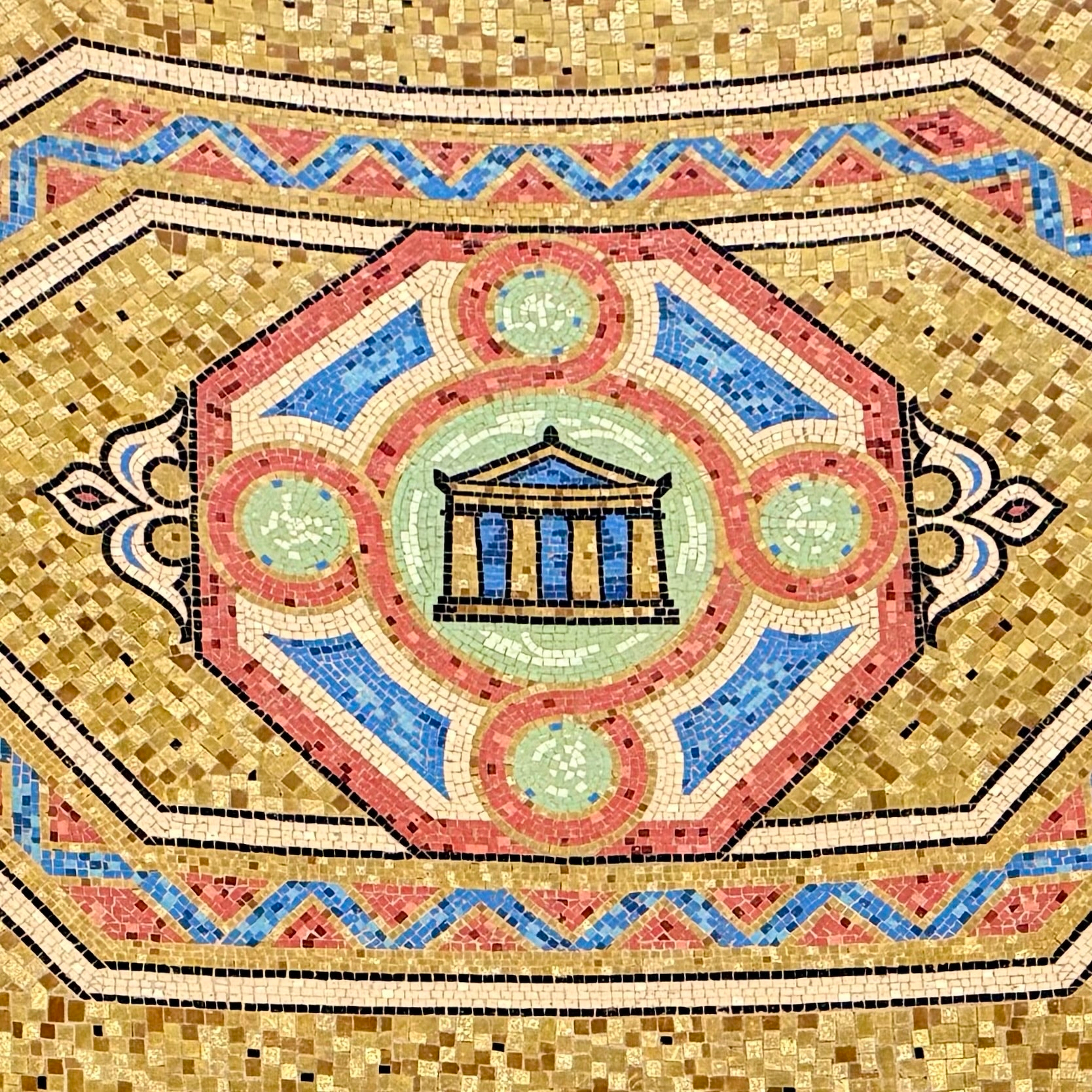 Mosaic artwork featuring a geometric pattern with an octagonal central design. In the center is an image resembling a classical Greek temple with columns, depicted in blue and gold. Surrounding the temple motif are concentric designs in green, pink, and blue. The background consists of small golden tiles, and there are intricate border patterns in pink, blue, and gold.