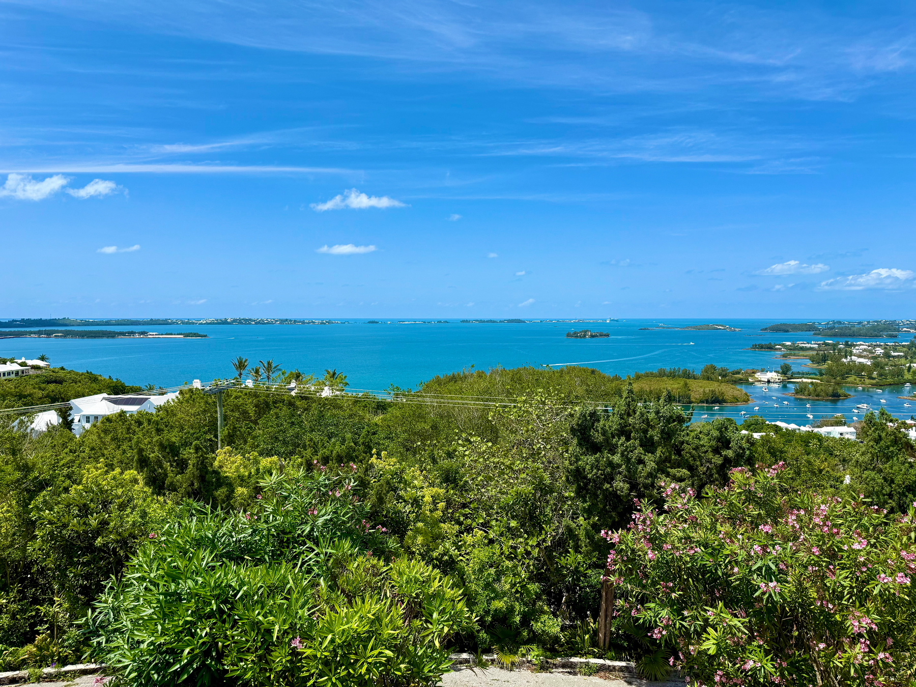A scenic coastal view under a clear blue sky with a few scattered clouds. In the foreground, lush green vegetation and flowering shrubs with pink blossoms frame the scene. Beyond the greenery, there are white-roofed houses partially visible among the trees. The midground features a calm turquoise sea dotted with small islands and peninsulas, and a few boats scattered across the water. In the distance, a thin strip of land stretches across the horizon, blending with the blue ocean and sky.