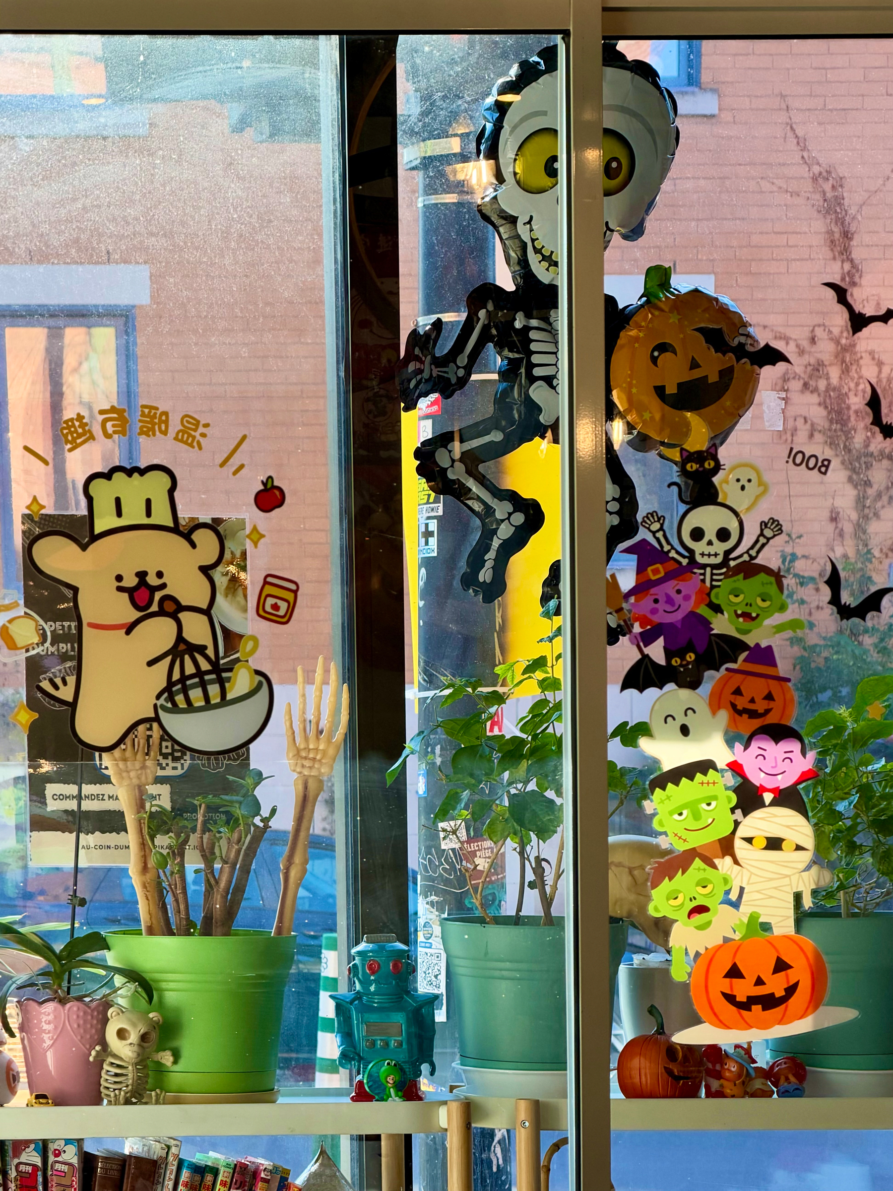 A window decorated for Halloween with various colourful decorations. On the left side, a cartoon bear wearing a chef hat is holding a whisk and a bowl, with small graphics like stars and an apple around it. Behind the window, two green potted plants have skeleton hands sticking up from the soil. A small turquoise toy robot and a tiny skeleton figurine sit on the windowsill near a pink potted plant. On the right side, there are Halloween-themed decorations, including a smiling skeleton with large yellow eyes, a pumpkin balloon with a cat on top, and a vertical stack of cartoon characters: ghosts, bats, Dracula, Frankenstein’s monster, a mummy, and a pumpkin. Additional small pumpkins and figurines are also on the windowsill. The outside view shows a brick building and reflections in the glass.