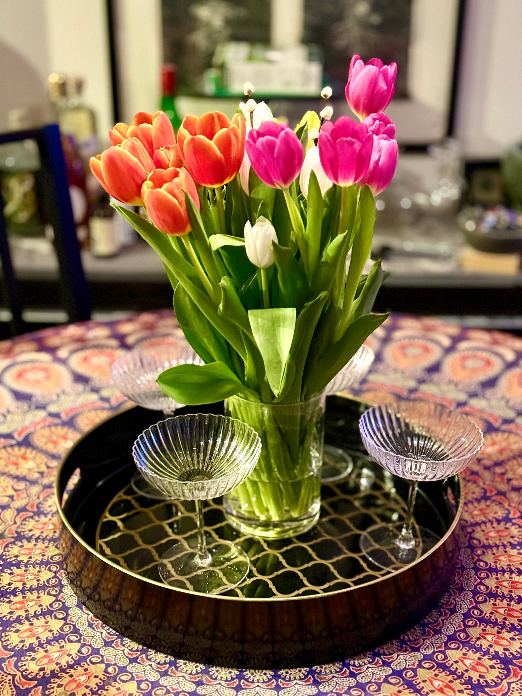 A bouquet of vibrant tulips in shades of red, pink, and white arranged in a clear vase. The vase is placed on a decorative round tray with a geometric pattern. The tray holds four empty, decorative glass dishes. Everything is set upon a table covered with a colorful, patterned tablecloth. The background is slightly blurred, showing various kitchen items.