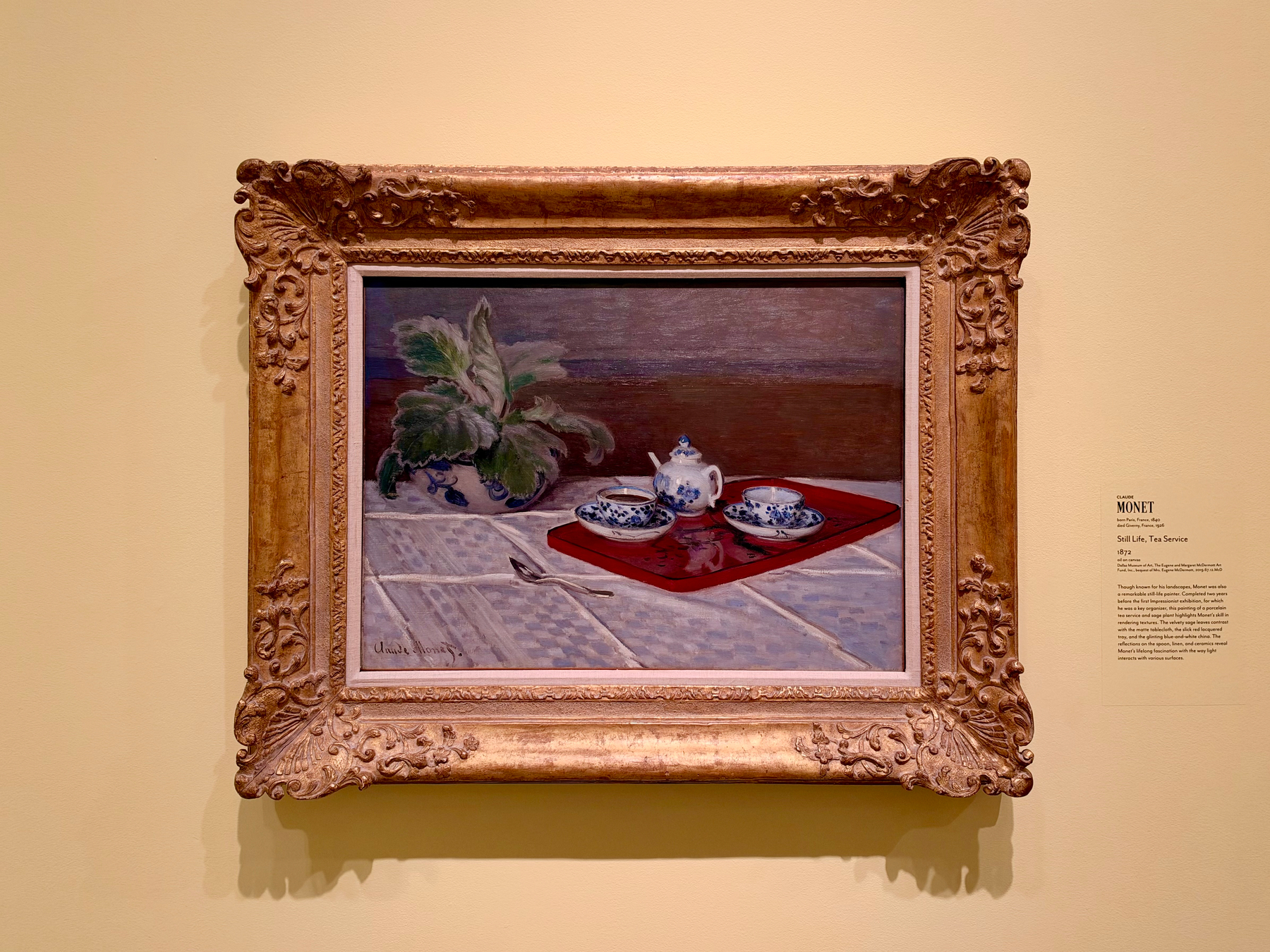 A framed painting hangs on a cream-coloured wall. The ornate golden frame features intricate floral carvings, particularly in the corners and along the edges. The painting depicts a still life scene with a white and blue porcelain tea set on a bright red tray, placed on a white tablecloth with a subtle pattern. Behind the tea set, a potted plant with large green and white leaves is positioned against a dark brown and purple background, giving depth to the composition. The tray contains a teapot and two cups with saucers, and a silver spoon lies diagonally across the tablecloth in front of the tray. A museum label to the right of the painting reads: “MONET. Still Life, Tea Service. 1872.” A short description below mentions Monet’s focus on light and surfaces in his work.