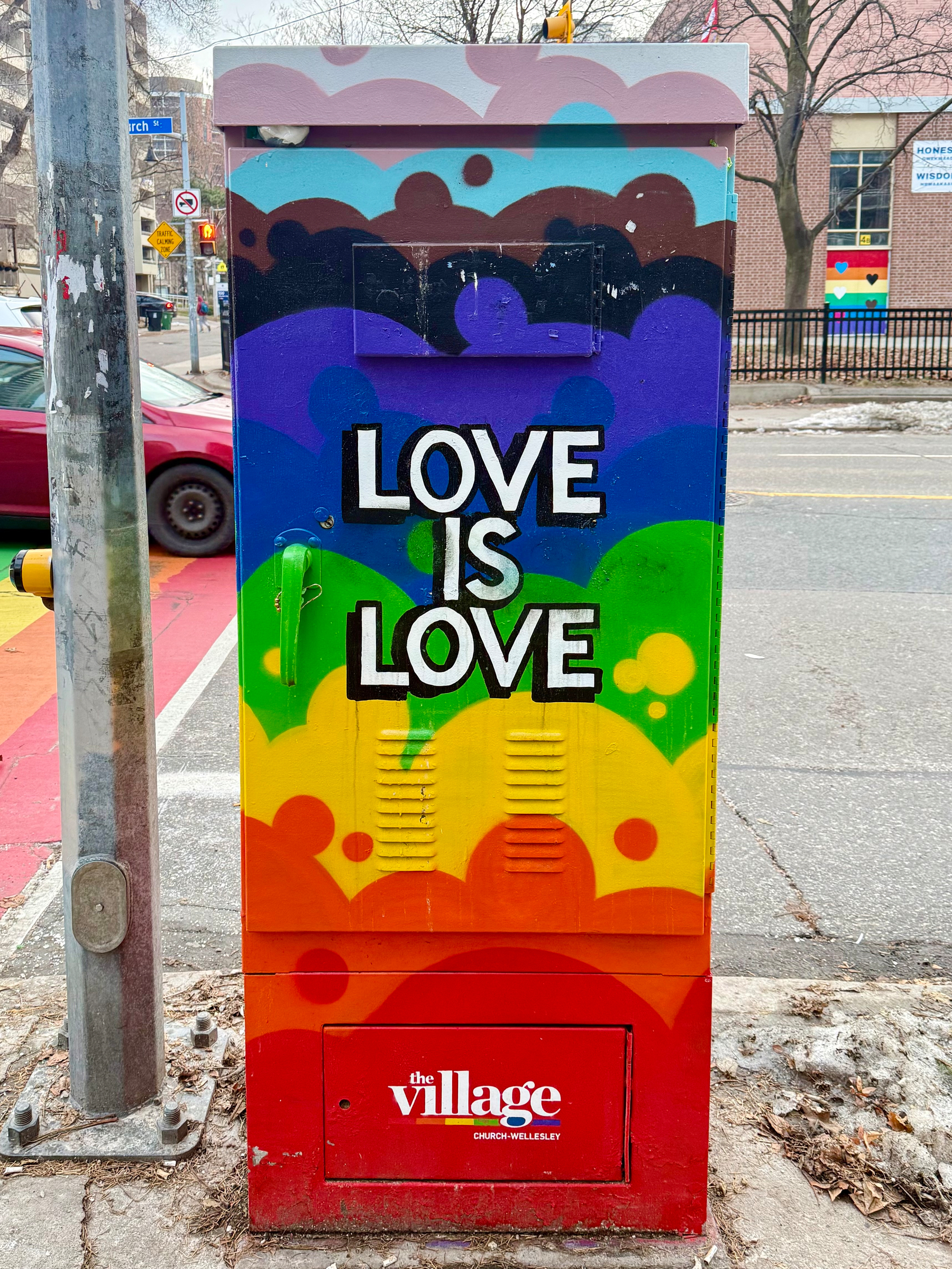 A street utility box is painted with a vibrant, rainbow-colored mural. The phrase "Love is Love" is prominently displayed in bold, white letters. Below, it reads "the village: Church-Wellesley" in smaller text. The background features colorful abstract shapes. The scene is set on a street, with a red car and buildings visible in the background.