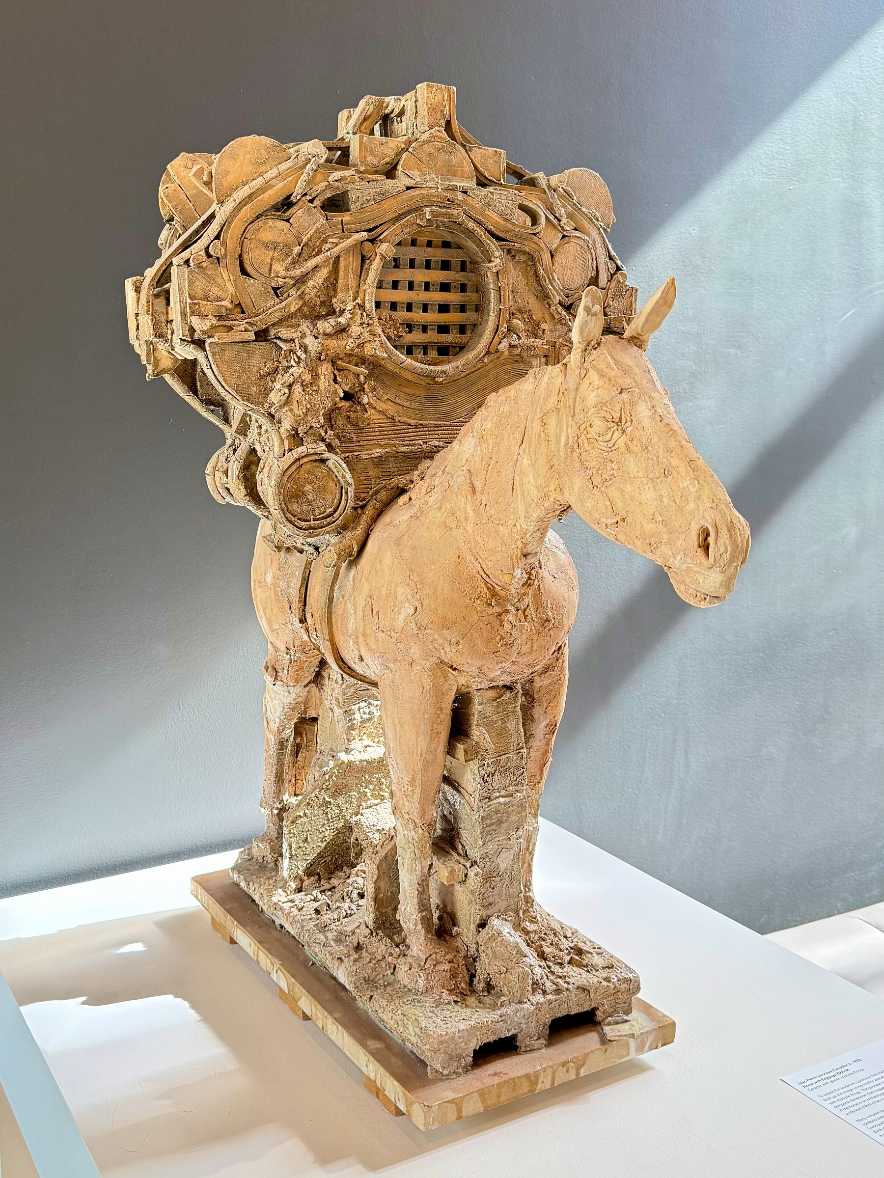 A sculpture of a horse made from a textured material, featuring intricate mechanical and abstract elements on its back. The horse stands on a rectangular base, under natural light against a gray background.