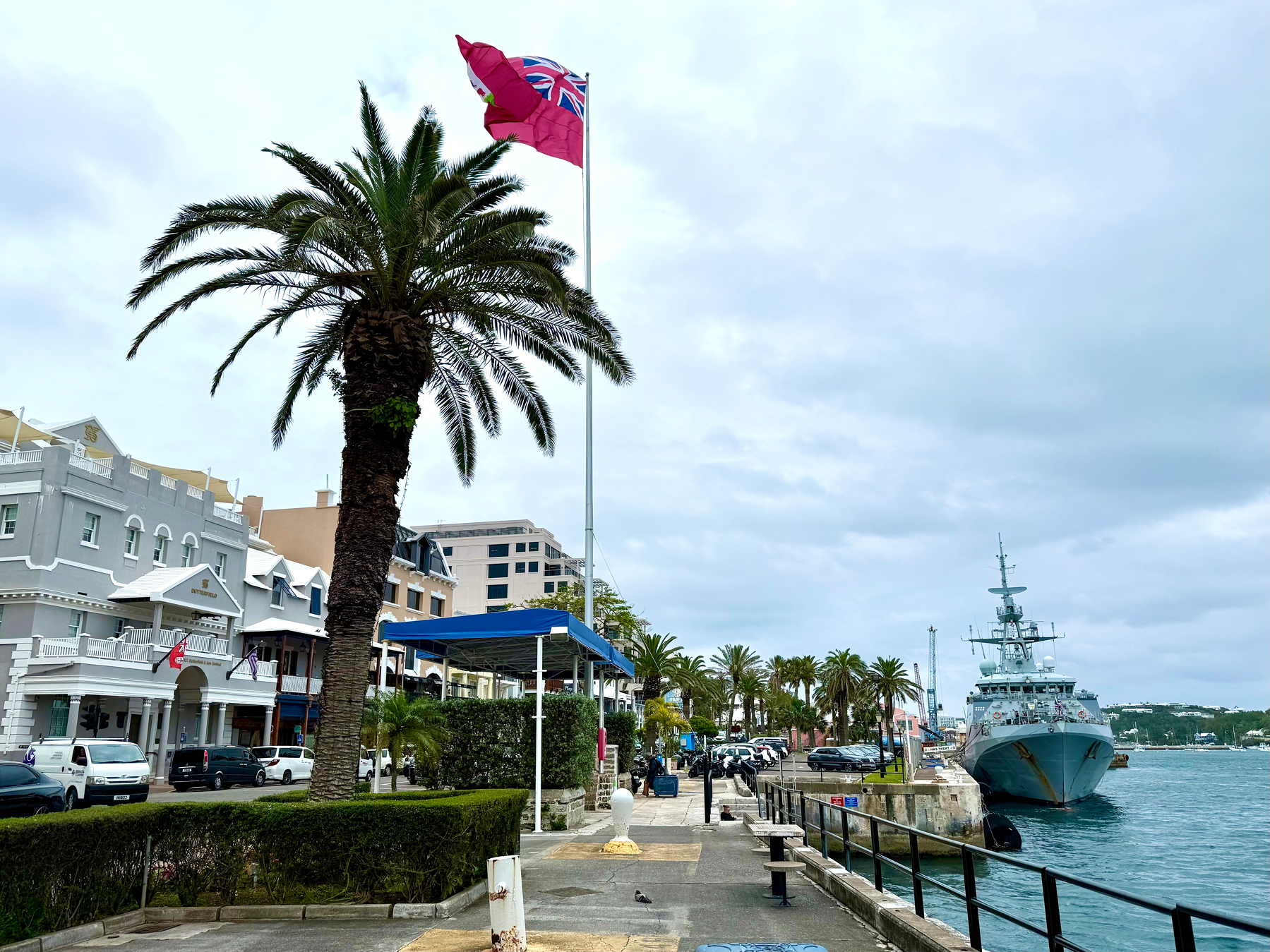 A coastal street scene shows a waterfront lined with palm trees and pastel-coloured buildings, including a grey structure with white trim and arched entrances. A large red flag featuring a Union Jack in the corner and a crest flutters on a tall pole near the water. A naval ship is docked along the stone seawall with its bow facing the camera, and several vehicles, including cars and scooters, are parked along the street. The sky is overcast, and the water appears calm with slight ripples. A pedestrian walkway with benches and railings runs alongside the water.