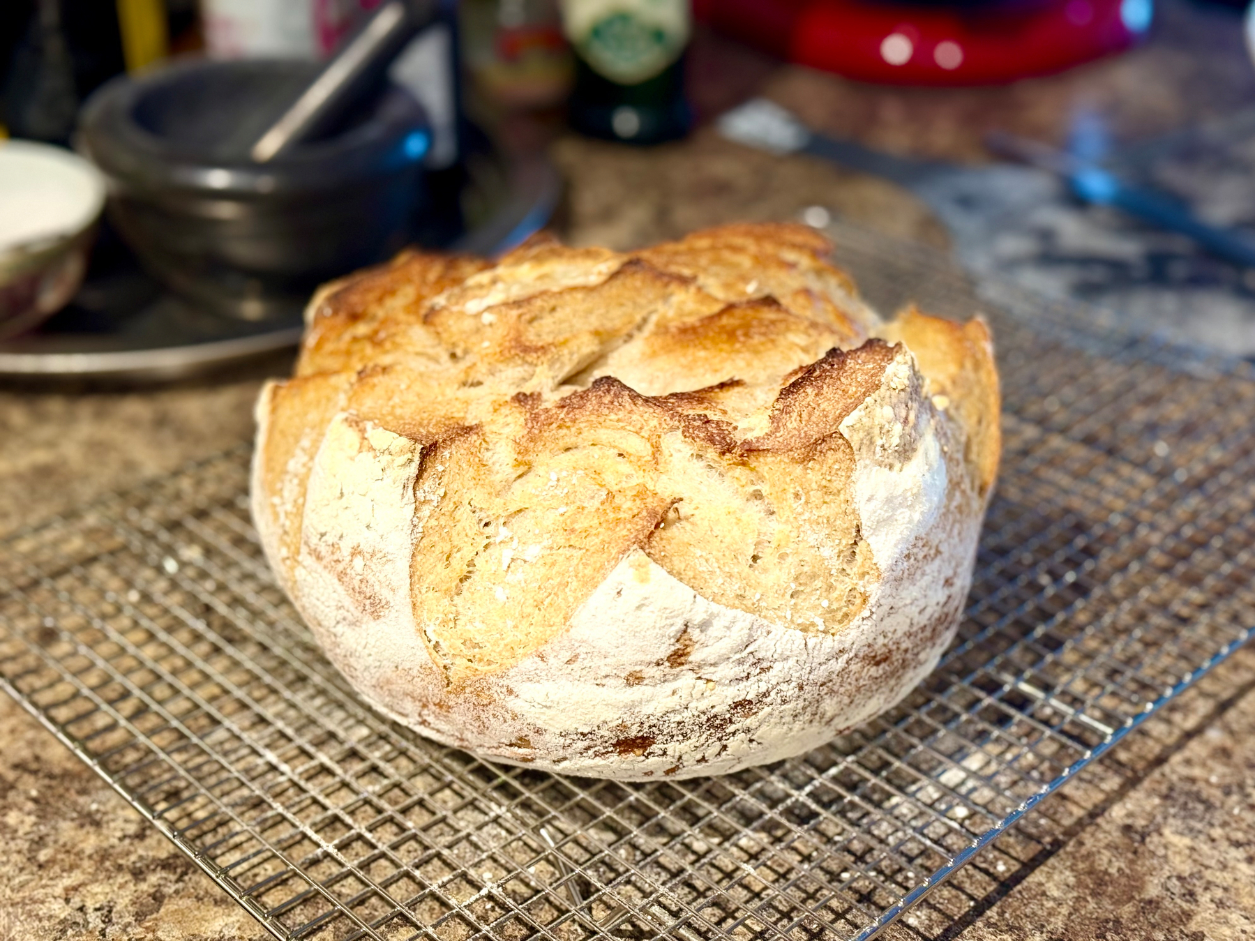 A freshly baked round loaf of rustic bread with a golden-brown crust and deep, jagged scoring sits on a metal cooling rack on a marbled kitchen counter. The crust has a slightly cracked appearance with areas dusted in flour. In the background, there is a black mortar and pestle, a white bowl with a floral rim, a green glass bottle, and a red kitchen appliance, all slightly out of focus.