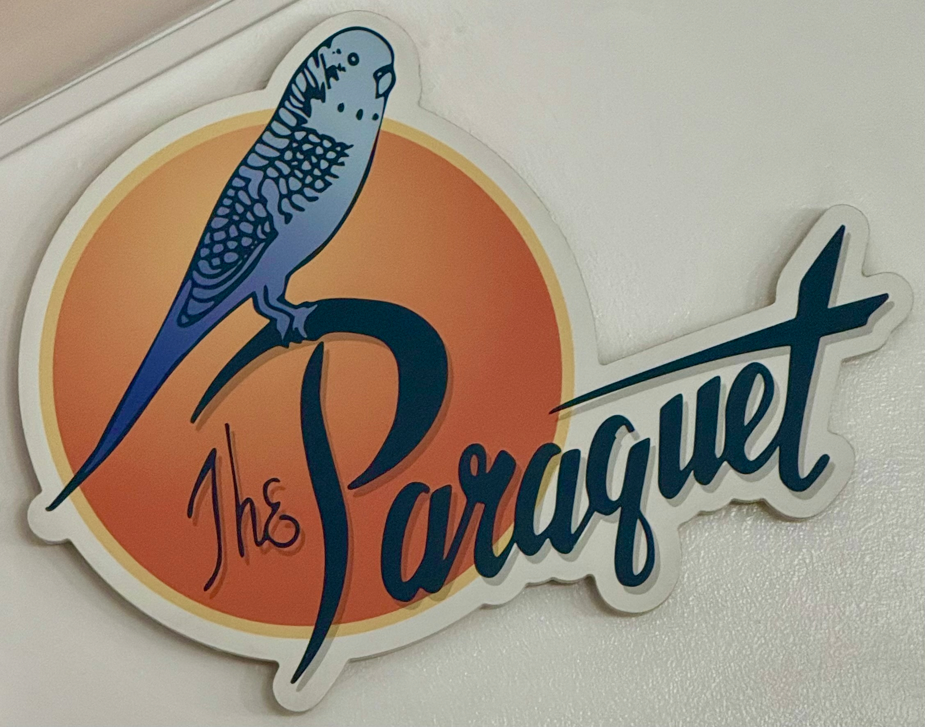 A logo featuring a blue bird perched on stylized text that reads The Parquet against an orange circular background. A decorative sign featuring a blue parakeet perched on the stylized text “The Paraquet.” The parakeet has a detailed feather pattern with dark blue markings on its wings and head. Behind the bird and text is a circular background with an orange gradient, creating a sunset-like effect. The overall design has a playful and retro aesthetic, with the text in a bold, flowing script. The sign is mounted on a white wall.