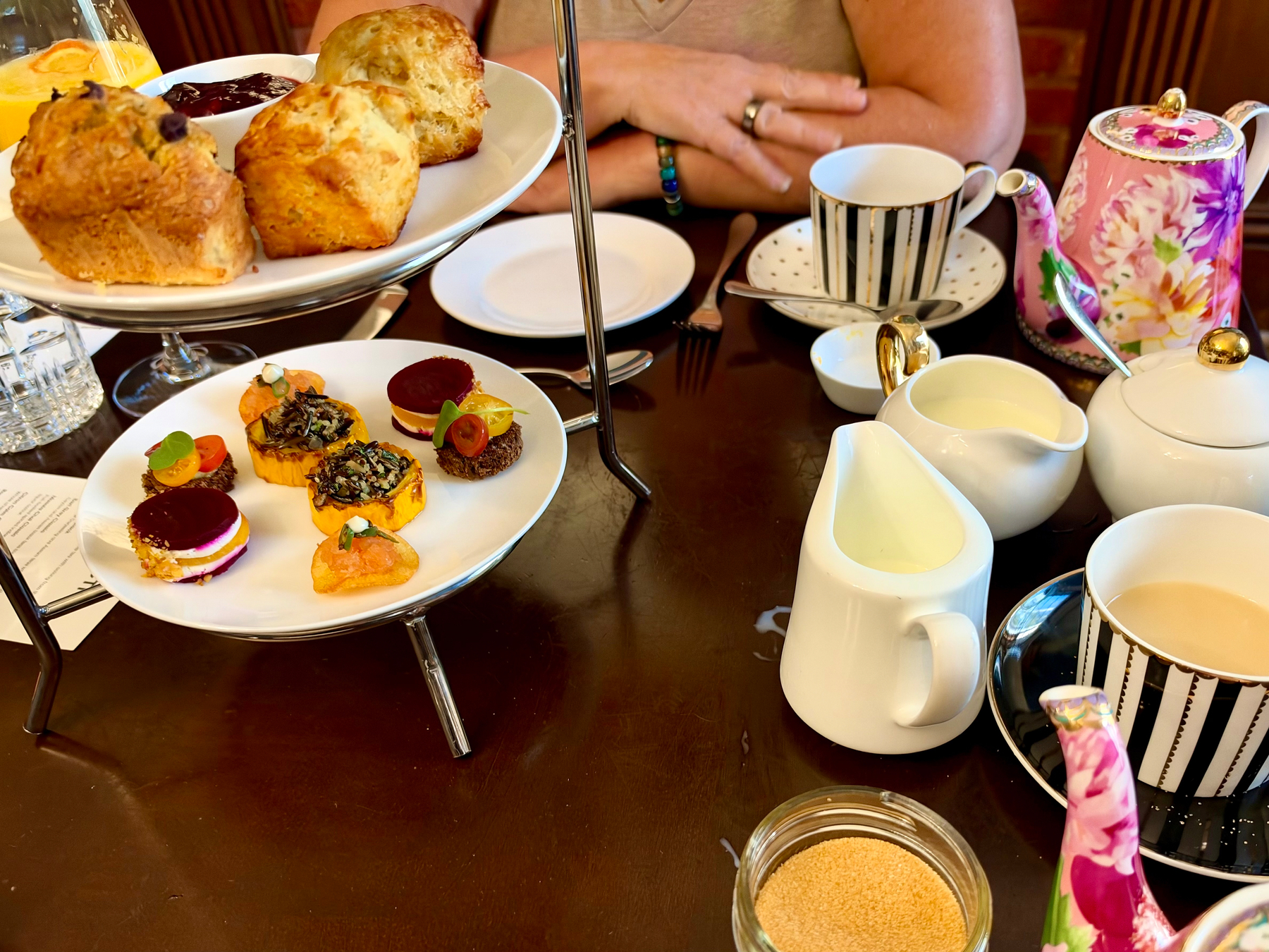 A table set for afternoon tea with various treats and drinks. A two-tiered serving tray holds golden scones on the top plate and an assortment of colourful bite-sized canapés on the lower plate, including layered beet and cheese stacks, small tarts, and garnished savoury bites. A black-and-white striped teacup with a matching saucer sits beside a small dessert plate, with a person’s folded arms visible behind it. A floral pink teapot with gold accents and a lid rests on the right, next to white ceramic creamers, a sugar bowl, and a small glass jar of brown sugar. A cup of tea with milk and a black-and-white striped saucer is nearby. The dark wooden table is set with cutlery, a glass of water, and an orange drink in the corner.