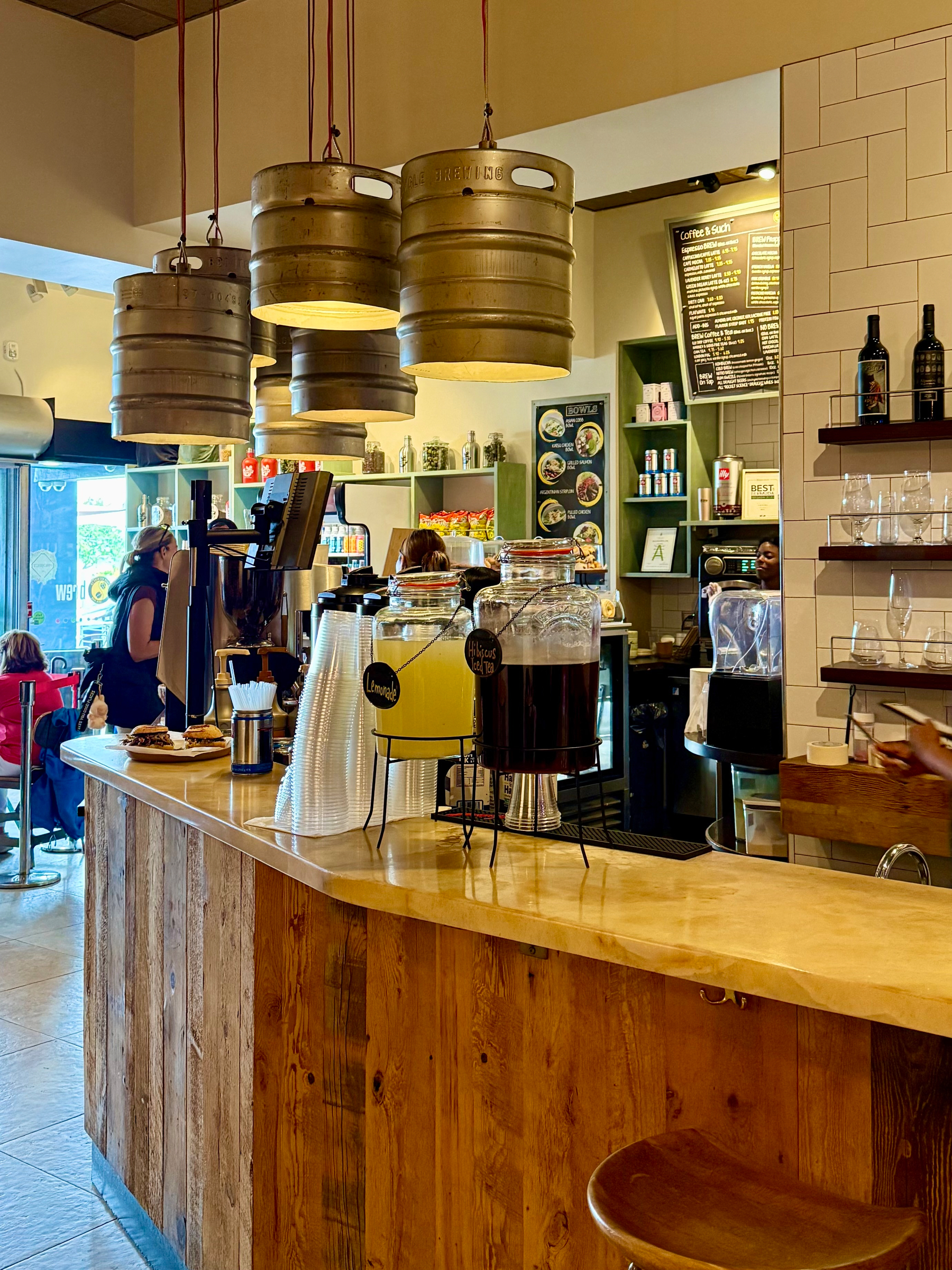 A cozy café interior featuring a rustic wooden counter with a polished stone top. On the counter, there are two large clear beverage dispensers, one filled with yellow lemonade and the other with dark hibiscus tea, each labeled with small black signs. Stacks of clear plastic cups sit nearby. Above the counter, four large metal beer kegs have been repurposed as hanging pendant lights with red cords. Behind the counter, baristas are preparing drinks, and shelves display jars, canned drinks, and small décor items. A black menu board with white and yellow text hangs on the wall, listing coffee and sushi offerings. On the right side, a wall with white tile holds shelves with bottles of wine and rows of glasses. In the background, customers are seated by the window, with natural light coming through the café’s glass doors.