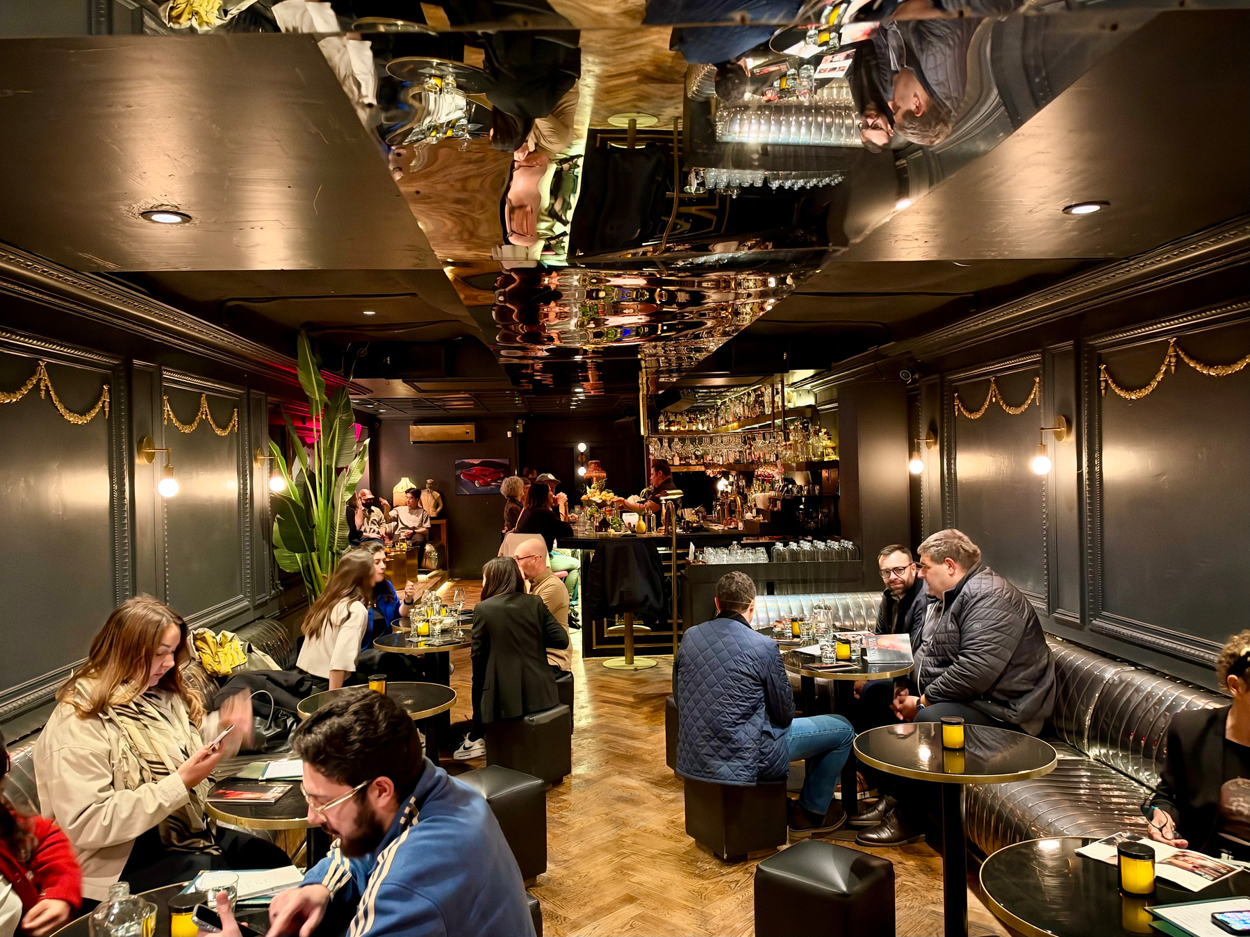 A cozy, dimly lit cocktail lounge with dark grey walls decorated with gold garlands and wall-mounted round lights. The space features a mirrored ceiling reflecting the patrons and décor below. Several groups of people are seated at small round tables with gold accents and black stools or a long metallic silver banquette. Drinks, menus, and candles are placed on the tables. In the foreground, a woman in a beige coat checks her phone, while a man in a blue jacket writes in a notebook. In the middle, a group of people engage in conversation, and in the background, bartenders work behind a well-stocked bar with bottles and glassware. Large potted plants add greenery to the modern, intimate space.