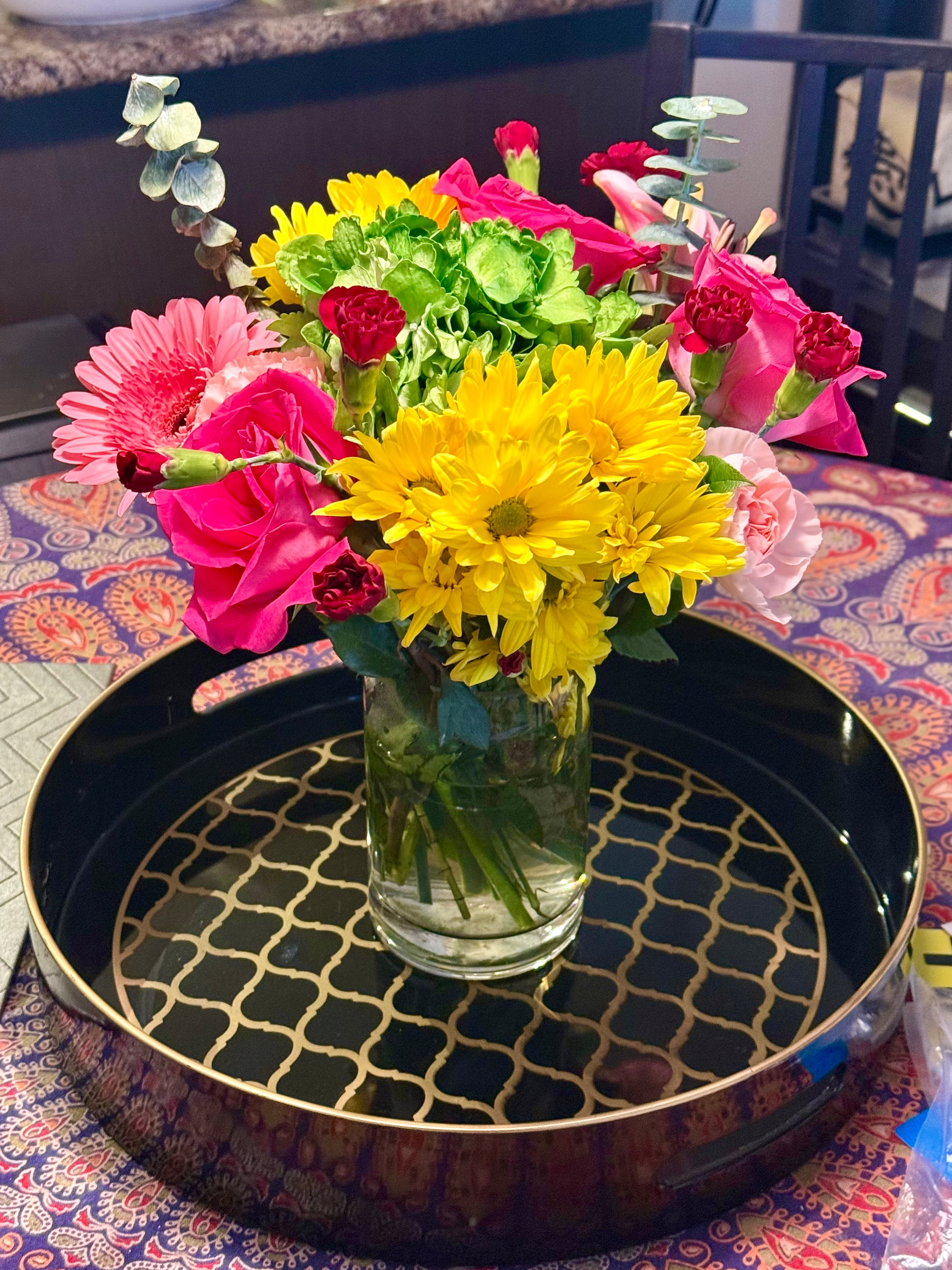 A glass vase filled with a vibrant bouquet of flowers sits on a round black tray with a gold lattice pattern. The bouquet includes bright yellow daisies, pink roses, pink gerbera daisies, small red carnations, light pink carnations, green hydrangeas, and sprigs of eucalyptus. The vase is placed on a patterned tablecloth with a detailed paisley design in shades of purple, orange, and red. In the background, part of a dark wooden chair and a granite countertop are visible, along with some blurred household items.