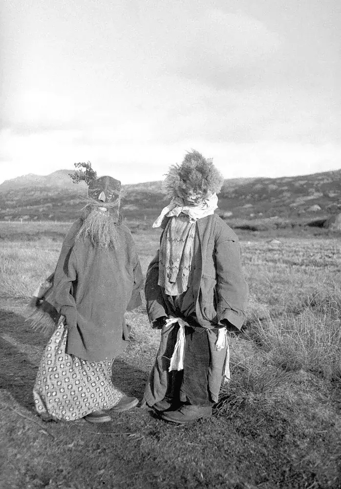 Black-and-white photograph of two people in costumed disguises standing in an open, grassy field with dry tufts of grass. Rolling hills rise in the background under a pale, overcast sky, and a low building or mound appears at the far right edge. The figure on the left wears a carved wooden mask with a large, pointed nose and a sprig of leaves tucked on top, plus a long, shaggy beard of straw or fiber. Its body is covered by a long, dark, loose coat, and it wears a patterned wrap or skirt with a repeating diamond motif above dark shoes. The figure on the right has a head covered by a bushy, tufted mass of fur or hair with a rough face visible beneath, and a white cloth ruffled around the neck. It wears a long dark coat with wide sleeves, a patterned cloth draped over the chest, and a white sash tied at the waist with ends hanging down, along with dark trousers and heavy boots. Both figures stand side by side, facing slightly away from the camera.