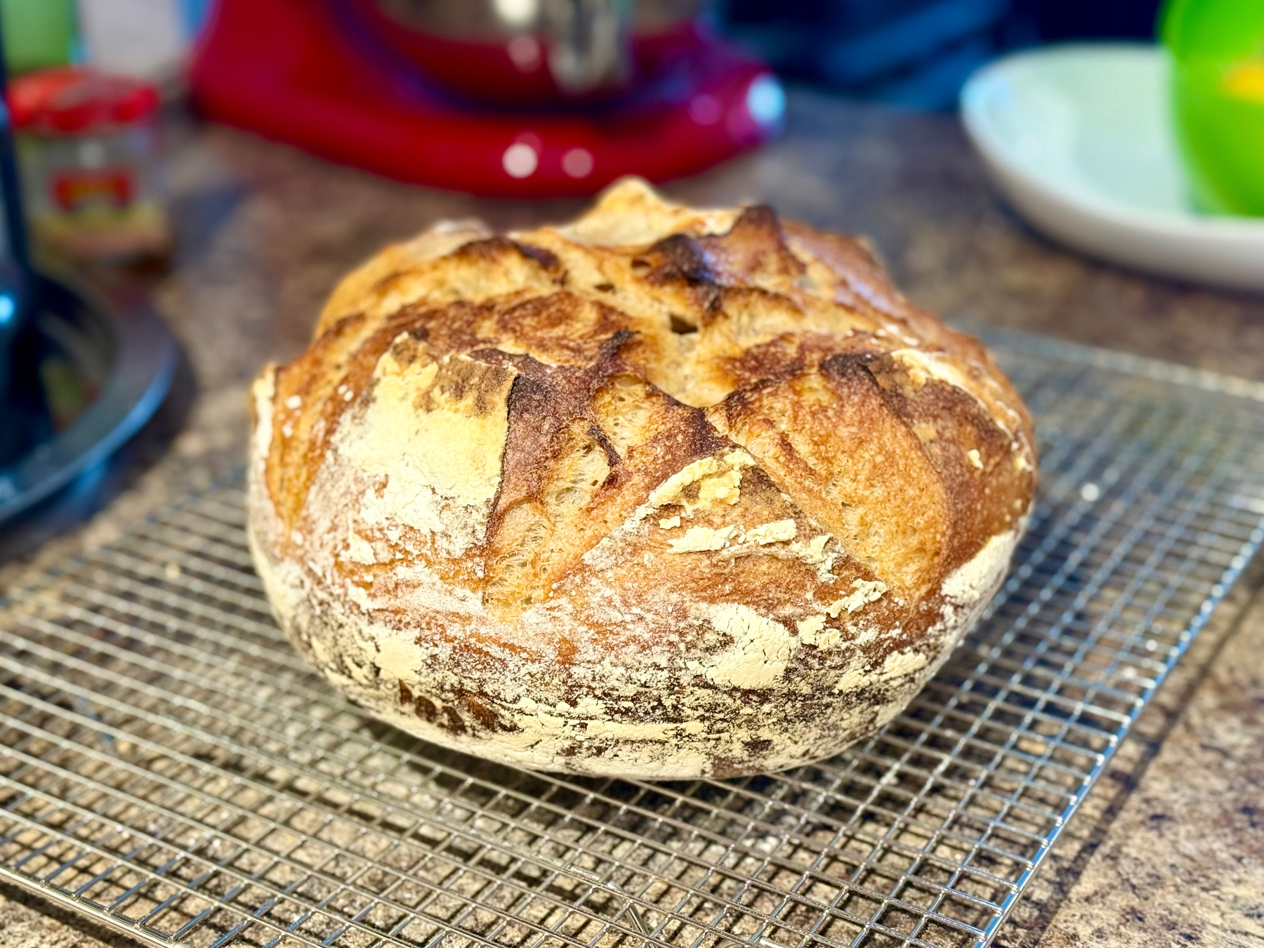 A freshly baked round loaf of artisan bread with a golden-brown crust sits on a wire cooling rack on a kitchen counter. The bread has a rustic appearance with a cracked, flour-dusted surface and deep scoring patterns on top. In the blurred background, there is a red stand mixer, a green bowl, a white dish, and a small jar on the counter. The scene is warmly lit, highlighting the texture and colour of the bread.