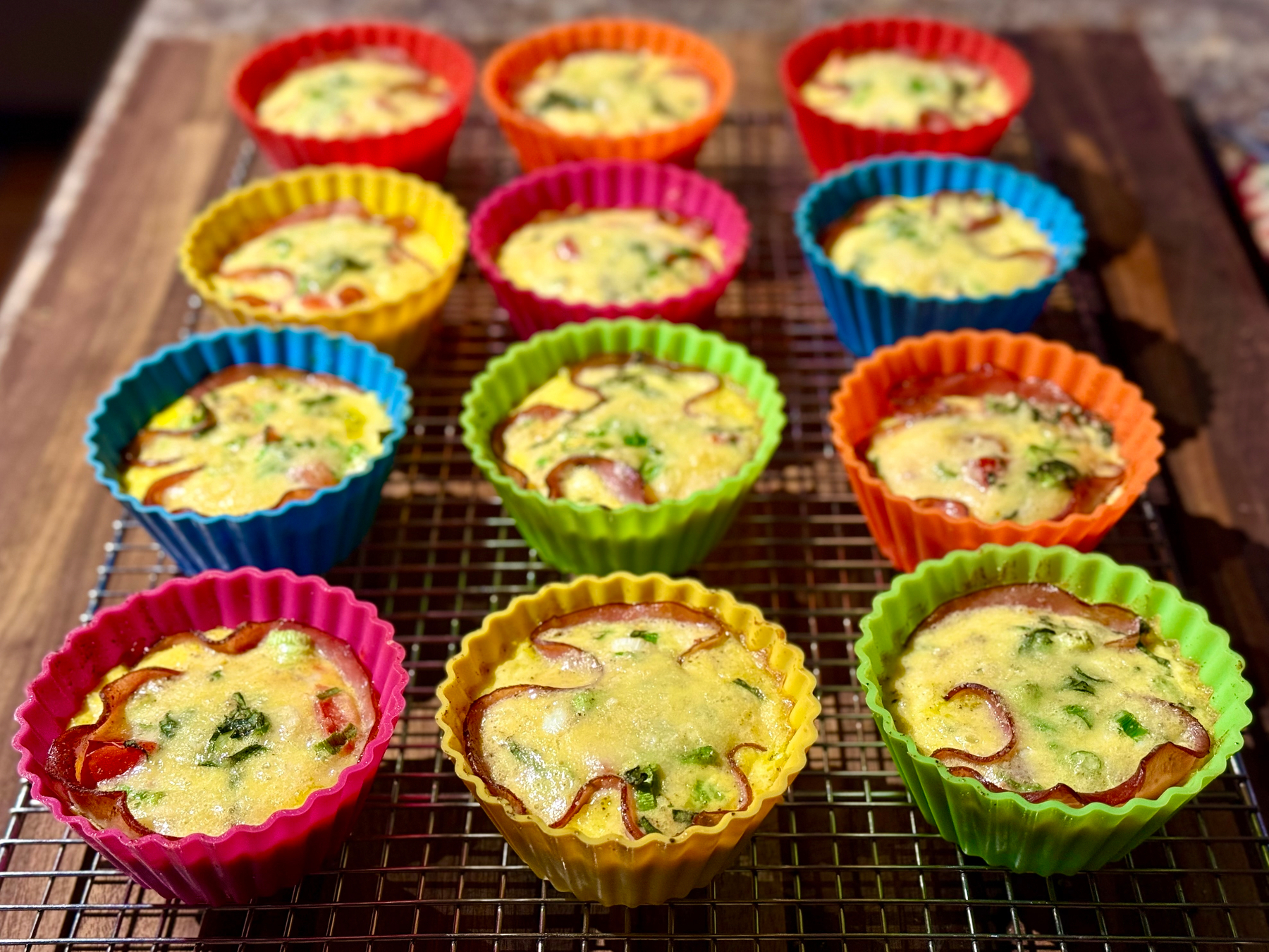 Colorful silicone muffin cups filled with freshly baked mini egg frittatas, placed on a cooling rack. Each frittata has visible ingredients like chopped vegetables. The muffin cups are in vibrant shades of red, orange, yellow, green, pink, and blue. The wooden countertop is partially visible.
