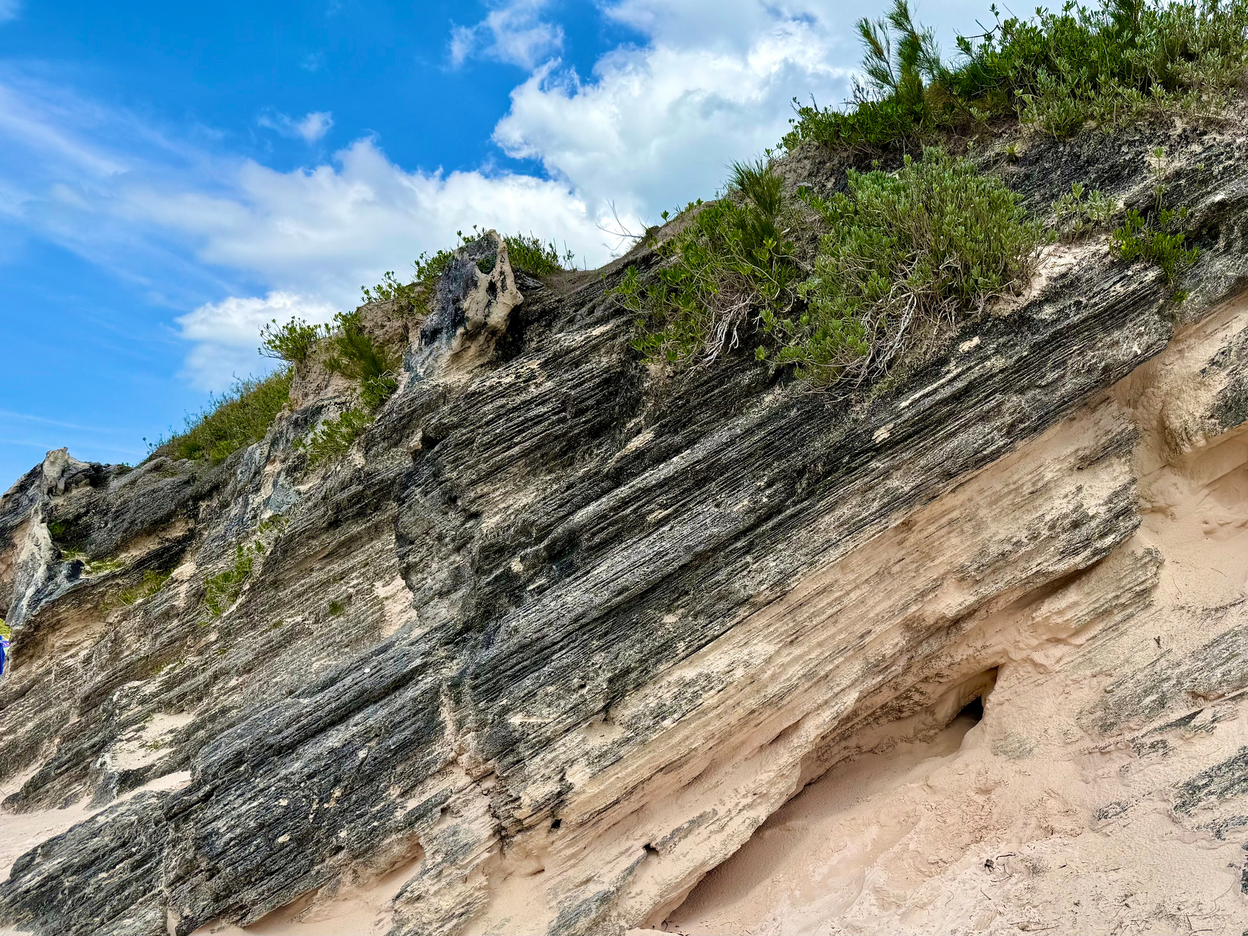 A steep coastal rock formation with layered, weathered stone and patches of light tan sand at the base. The rock has dark grey and black streaks running diagonally, with areas of erosion creating small holes and crevices. Green vegetation, including low shrubs and grasses, grows along the top edge of the rock, contrasting with the rough stone surface. Above, the sky is bright blue with scattered white clouds.