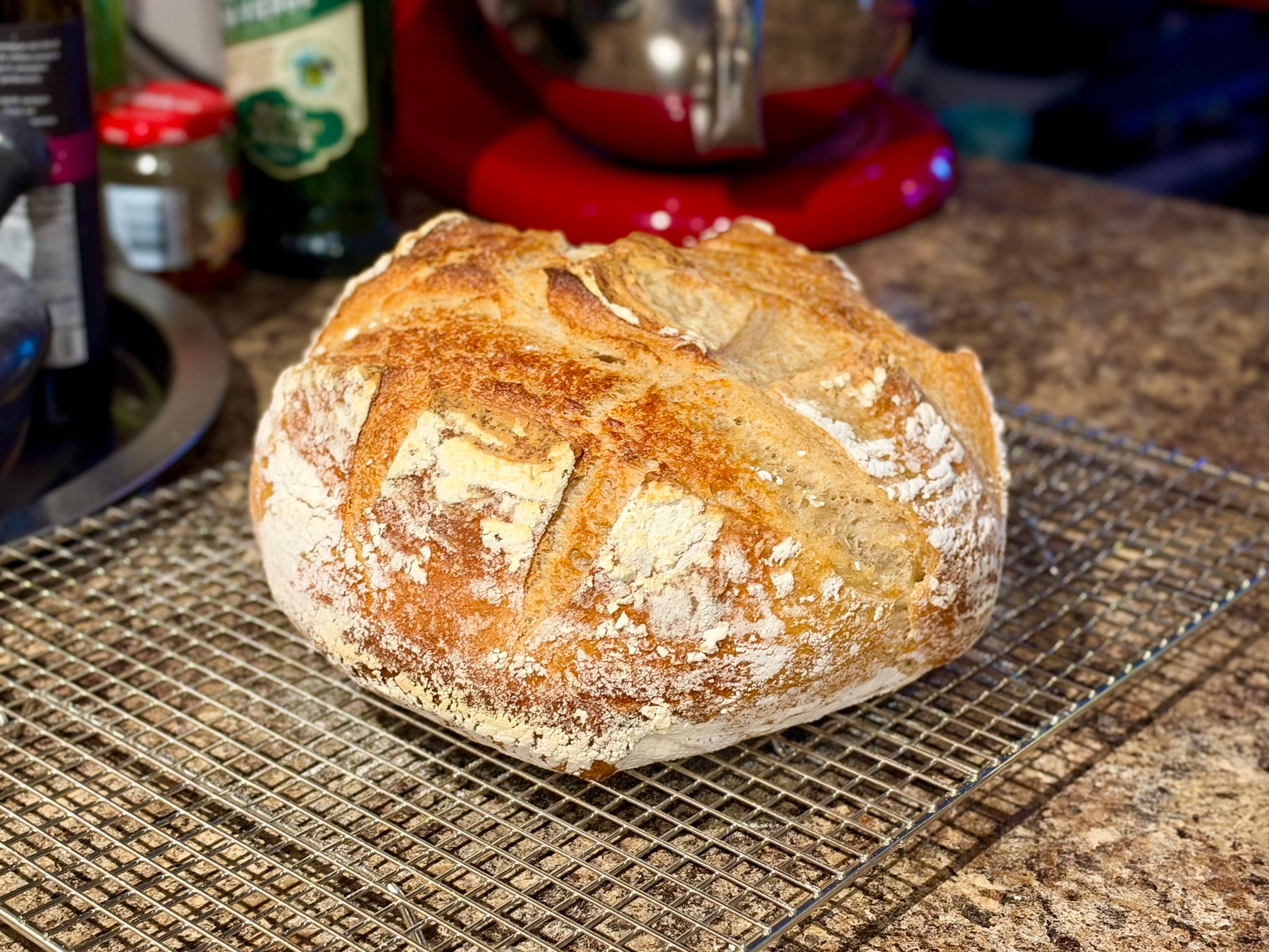 A freshly baked round loaf of bread with a golden-brown crust sits on a metal cooling rack on a speckled kitchen countertop. The bread has a rustic appearance with a dusting of flour and deep scoring patterns across the top, forming an “X” shape with additional diagonal cuts. The crust is textured and crispy, with some areas slightly darker from baking. In the background, a red stand mixer, various bottles, and jars are partially visible, slightly blurred to focus on the bread.