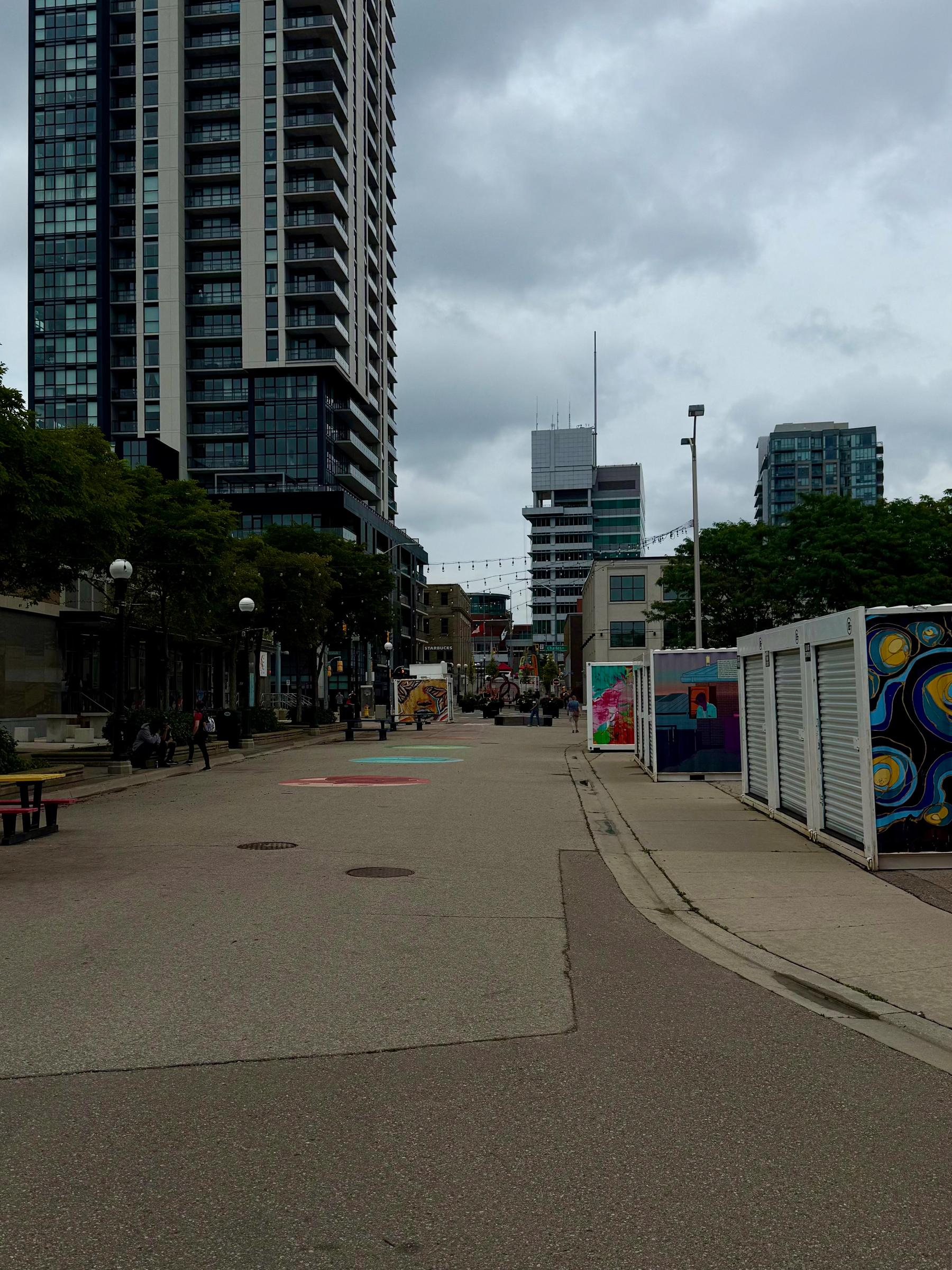 A wide pedestrian street is lined with trees, benches, colorful murals, and tall modern buildings under a cloudy sky.