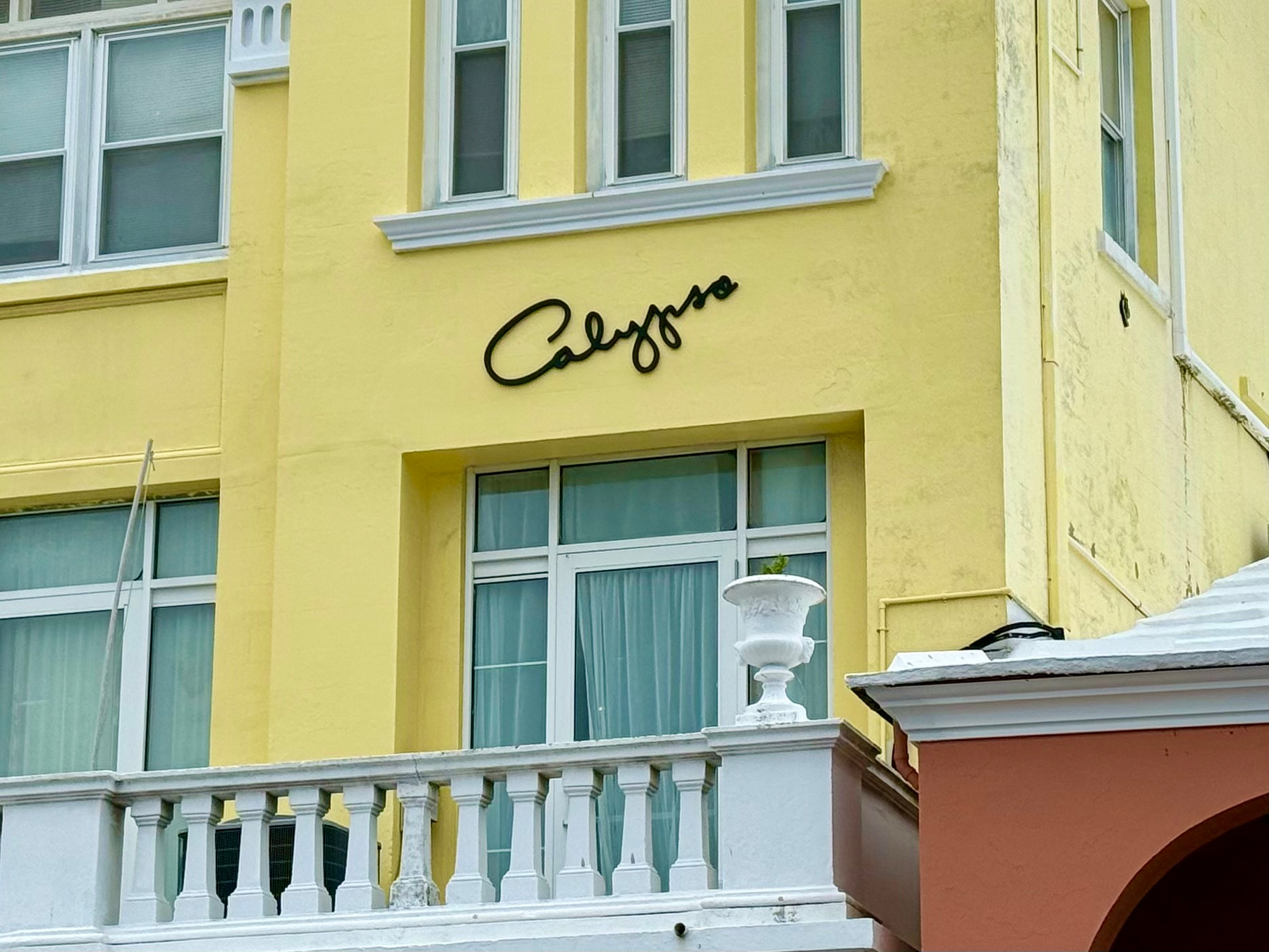 A yellow building with white trim and large windows is shown, featuring a white balcony with decorative balusters. A white urn planter sits on the balcony’s edge. Above the balcony, the word “Calypso” is displayed in black, cursive-style script mounted on the wall. Part of a peach-coloured building with a white roof is visible on the right side of the image.