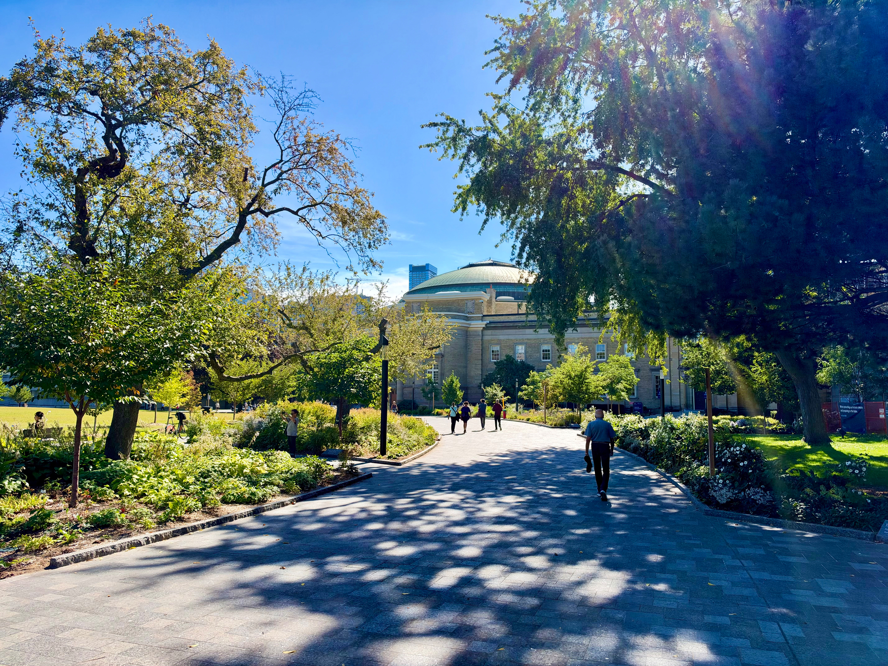 A sunny park scene with a wide paved pathway lined by lush green trees and landscaped flowerbeds. The pathway leads toward a large domed building in the background, with beige stone walls and arched windows. Sunlight filters through the trees, casting long shadows and light rays across the path. Several people are walking along the path, while others are sitting or standing on the grass to the left. The sky is clear and bright blue, and the overall atmosphere is peaceful and vibrant.
