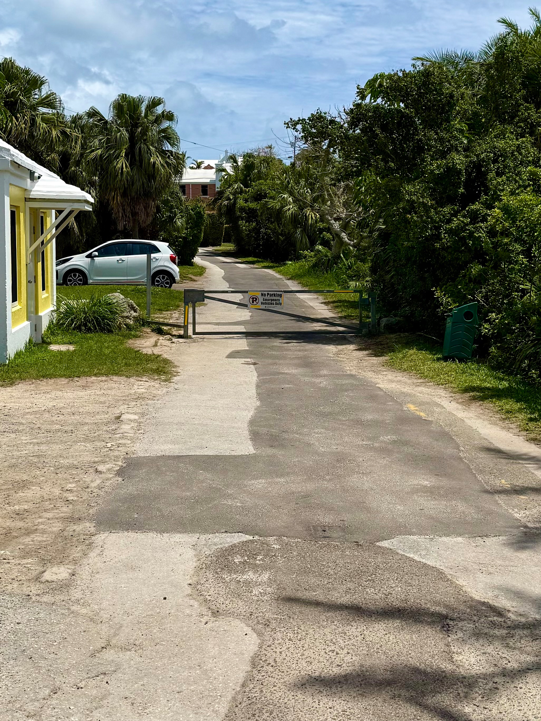 A narrow paved road flanked by greenery and a yellow building leads up to a barrier with a No Parking, Private Driveway sign and a silver car parked nearby.