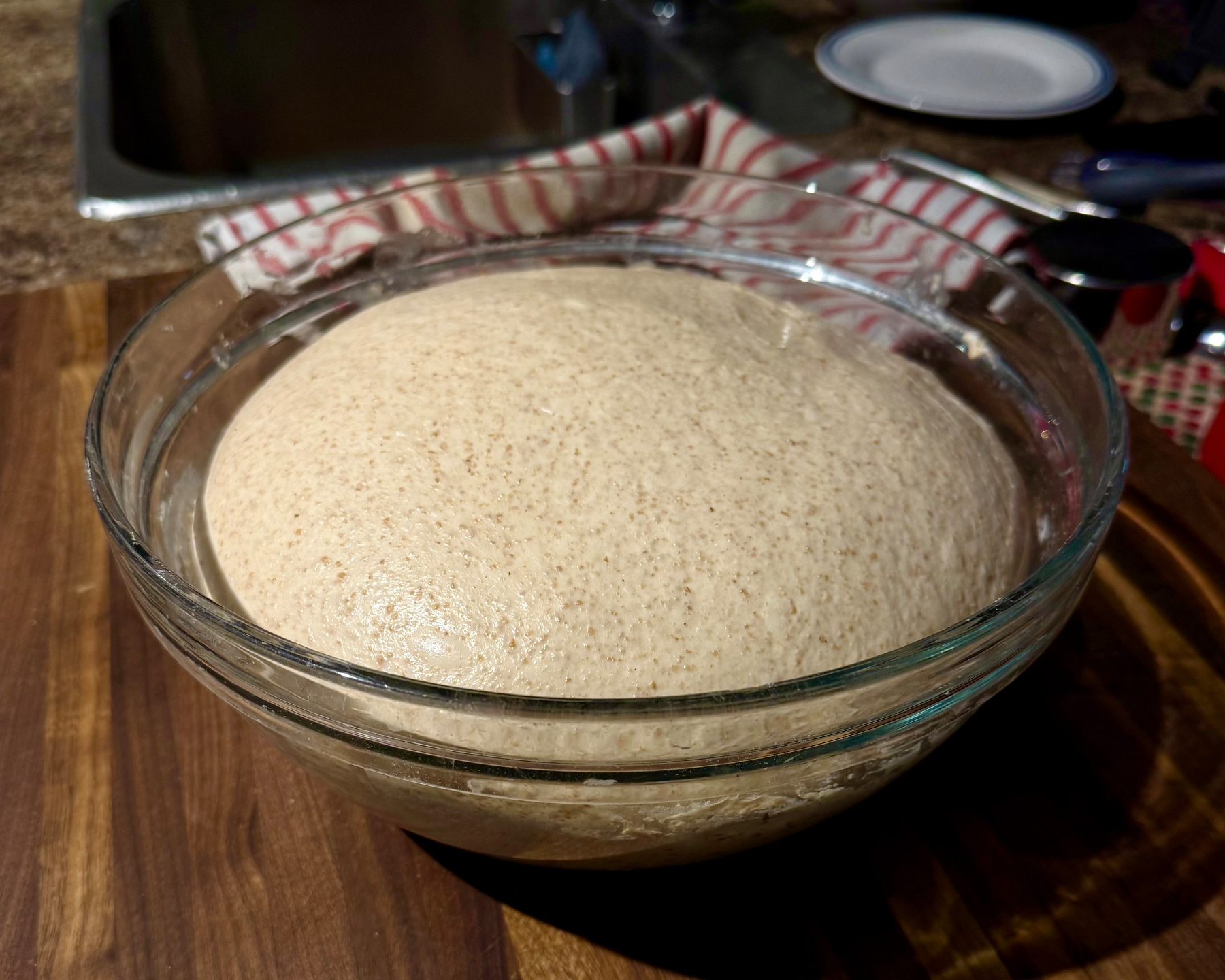 A glass bowl filled with risen bread dough sits on a wooden countertop. The dough has puffed up and is smooth with a slightly speckled texture, indicating whole grain flour. Behind the bowl, a red and white striped cloth is draped over the edge of the counter near a stainless steel sink. In the background, kitchen items including a white plate with a blue rim, a black-handled utensil, and measuring cups are visible, slightly out of focus.