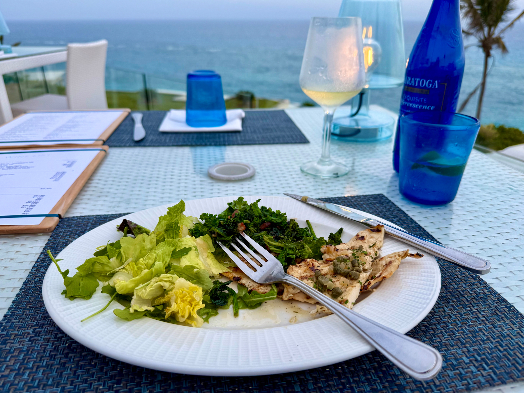 A seaside dining setup featuring a white wicker table with a woven blue placemat, holding a partially eaten meal. The plate has grilled fish topped with capers, sautéed greens, and a small leafy salad with romaine and mixed greens. A fork and knife rest on the plate. In the background, there is a glass of white wine, a blue bottle of sparkling water, a blue glass with a lime slice, and a blue glass lantern with a candle inside. The ocean and horizon are visible beyond the glass railing, with a palm tree to the right and white patio furniture around the table. Two menus in wooden frames lie on the table.
