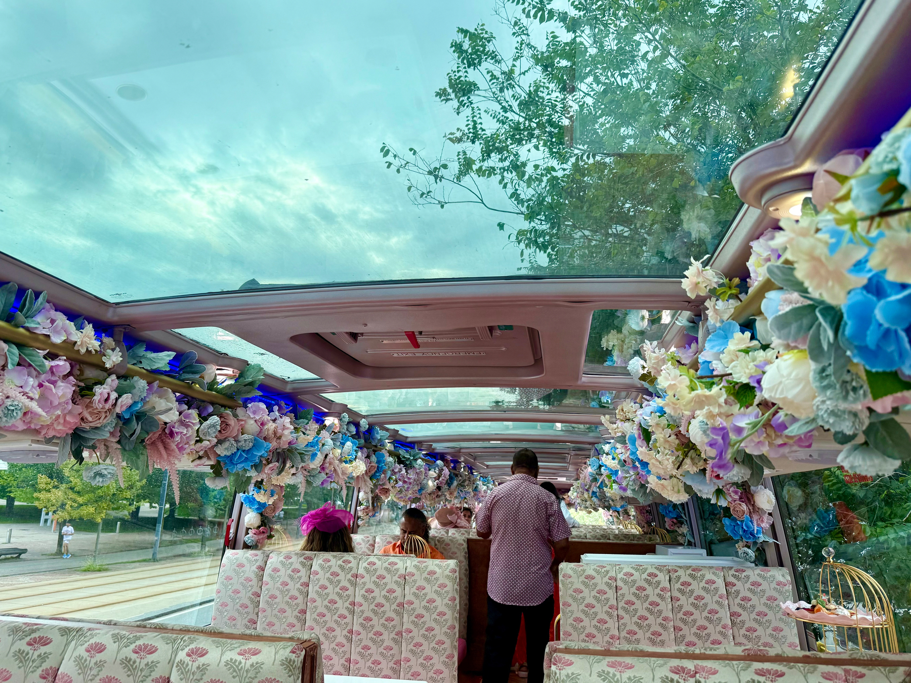 The photo shows the interior of a decorative double-decker bus or tram with a glass roof, allowing a view of the sky and tree branches above. The ceiling and upper walls are lined with colourful artificial flowers in shades of pink, purple, blue, and white, creating a festive and whimsical atmosphere. The seats are upholstered in cream fabric with a pink floral pattern. A few passengers are seated, and one person stands near the middle of the bus, facing forward. The right side shows a small golden birdcage-like stand holding some items. The setting appears bright and cheerful, with the outside view revealing a street with tram tracks, sparse greenery, and a pedestrian walking.