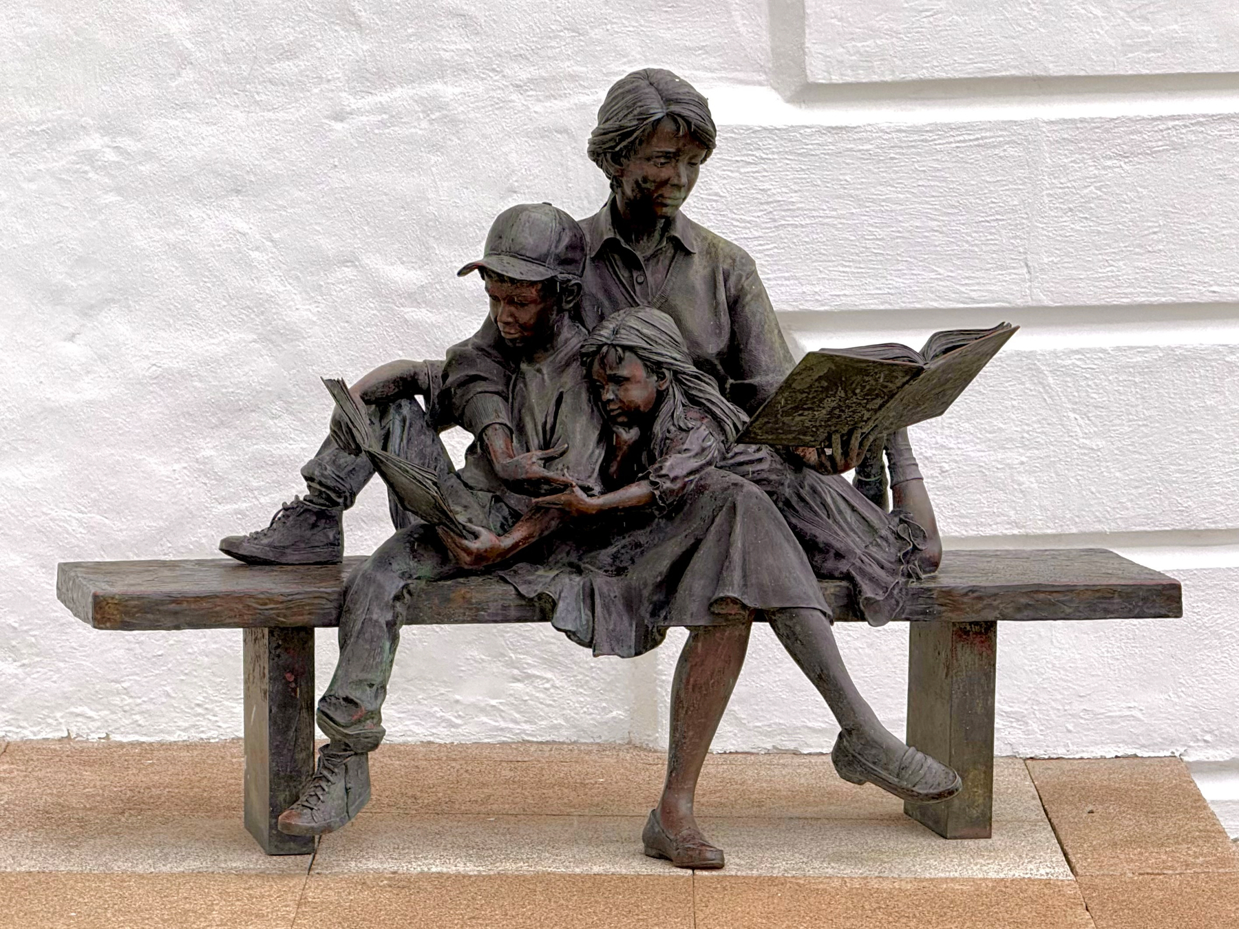 A life-sized bronze sculpture of three figures sitting on a bench against a white textured wall. The sculpture depicts an adult and two children reading together. The adult, seated in the centre with legs crossed, holds an open book while gazing down at it. A young girl sits close beside the adult, also looking intently at the book, her arm resting along the adult’s lap. A boy in a baseball cap sits on the other side, leaning forward with one knee up on the bench while reading a smaller book held in his hands. The bench is simple and rectangular, with the sculpture integrated into its surface. The ground beneath is paved with light brown tiles.