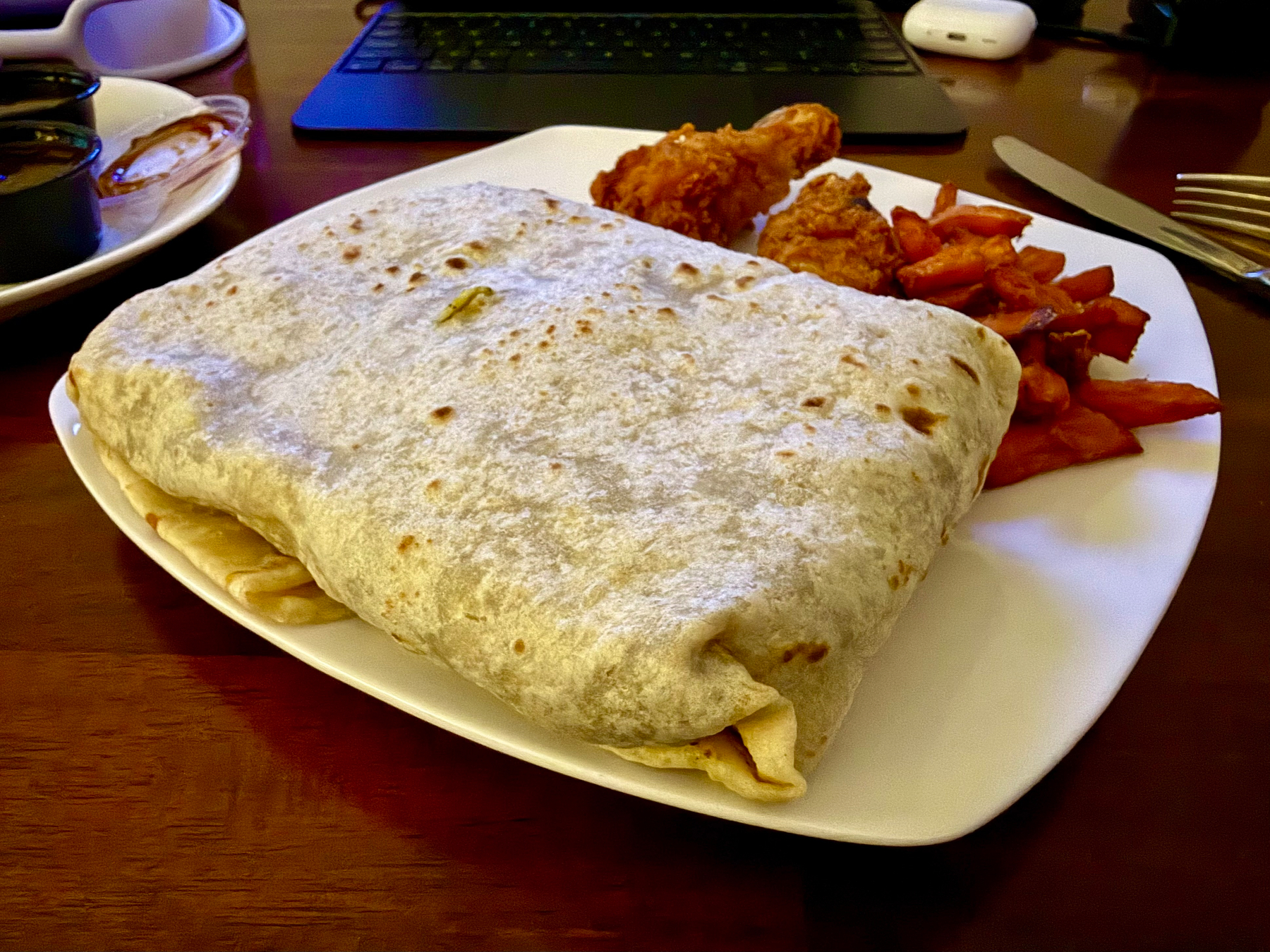 A square white plate holds a very large, rectangular roti. Behind the roti are two pieces of fried chicken and a small pile of reddish-orange sweet potato fries. To the left, a separate small white saucer contains two black dipping cups, one filled with a dark sauce and another lid with sauce residue. In the background, a tablet with a keyboard cover and a white wireless earbud case are visible on a wooden table, along with a fork and knife on the right side of the plate.