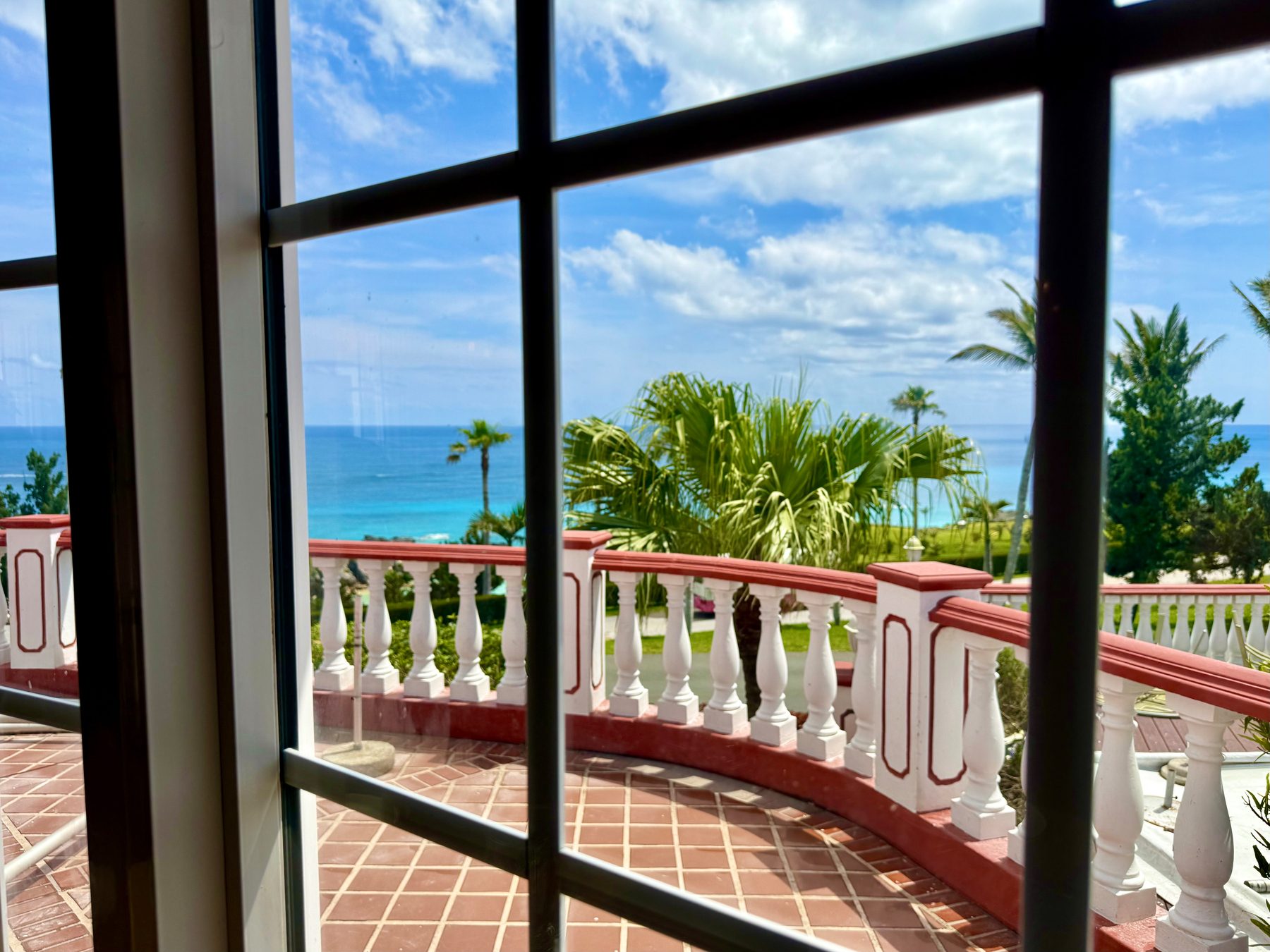 View through a window with white frames and black grid lines, showing a tiled terrace with a curved white and red balustrade. Beyond the terrace are lush green palm trees and other tropical foliage, with a bright turquoise ocean meeting the horizon under a partly cloudy blue sky. The scene conveys a coastal, tropical setting with vibrant colours and a serene atmosphere.