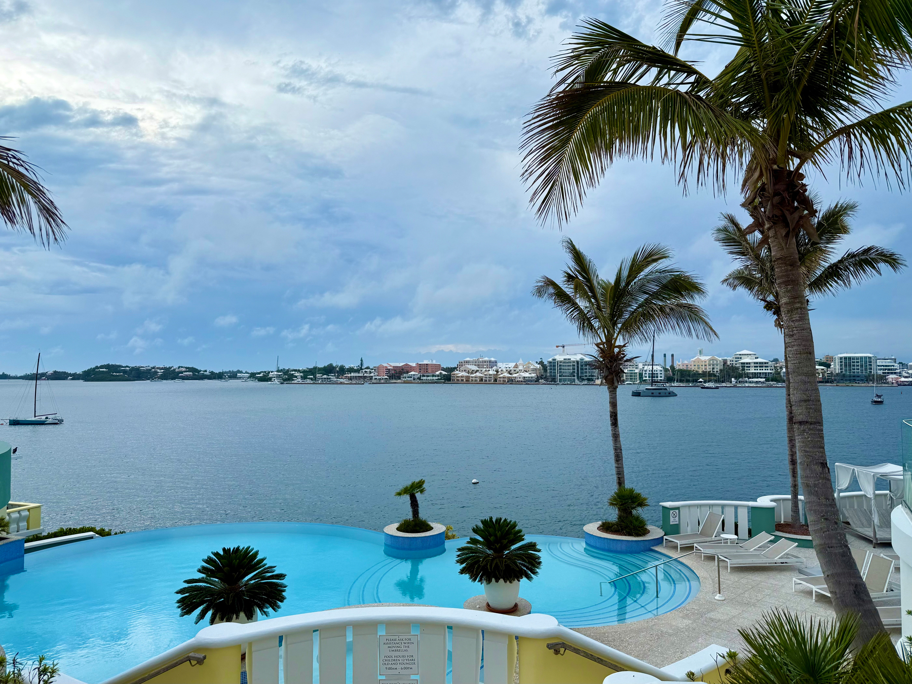 A serene waterfront scene featuring an infinity pool surrounded by potted tropical plants and tall palm trees. The pool’s bright blue water reflects the cloudy sky, and curved white railings frame its edges. Beyond the pool, a calm body of water stretches toward a distant shoreline lined with low-rise pastel-coloured and white buildings, as well as some modern structures. Several boats and sailboats float peacefully in the harbour. The sky is mostly overcast, with patches of lighter clouds suggesting a break in the weather.