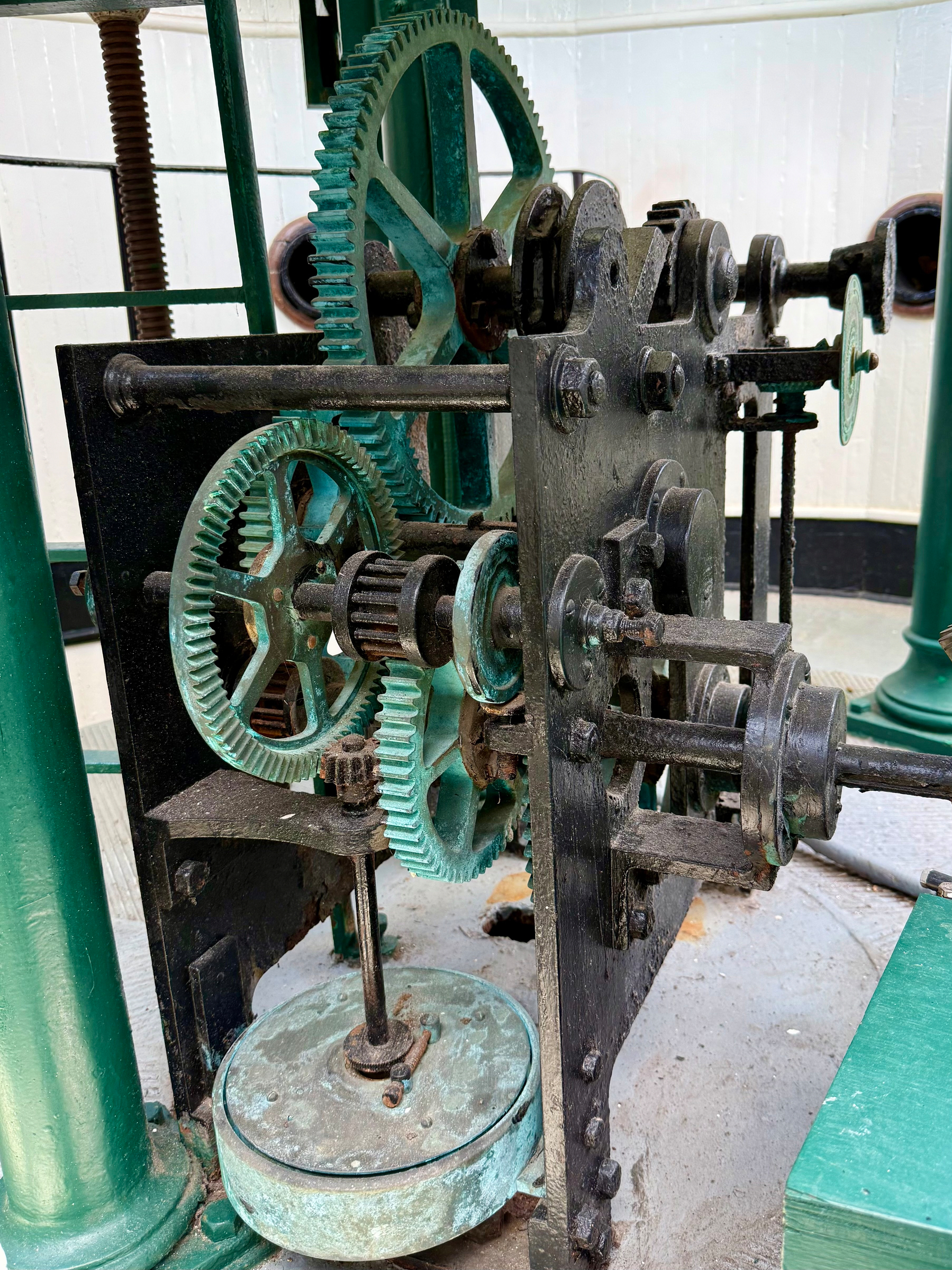 A close-up view of an antique mechanical clock movement with a series of interlocking gears and shafts. The gears are primarily green with signs of oxidation and wear, and some are mounted horizontally while others are vertical. The mechanism is made of metal, with a black iron frame supporting multiple rotating parts and large screws. A round, weathered metal weight sits at the base, connected to a vertical rod that engages with the gears above. The surrounding area includes green metal columns and a concrete floor, with a white, ribbed wall in the background. The entire mechanism has a rustic, aged appearance, showing signs of use and exposure to the elements.