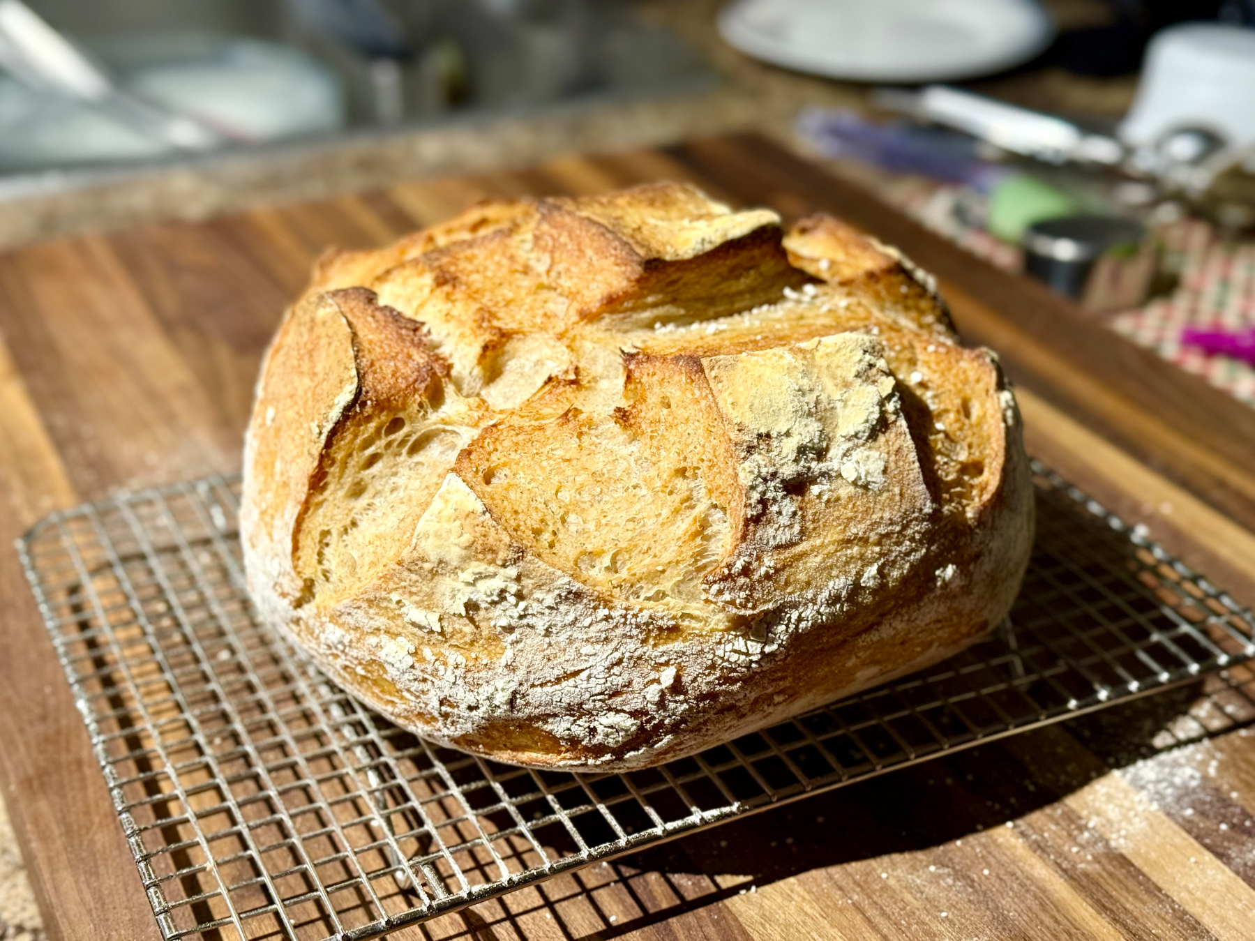 A freshly baked round loaf of artisan bread with a golden-brown crust sits on a metal cooling rack atop a wooden countertop. The bread has a rustic, crusty exterior with deep, irregular scoring patterns and a dusting of white flour on the surface. The loaf appears airy and well-risen, with cracks along the crust exposing the soft, slightly yellow interior. The background shows a blurred kitchen counter with utensils, a plate, and various items out of focus.