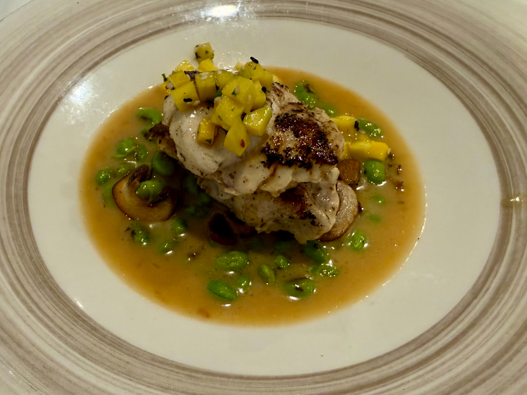A plated gourmet dish featuring a seared rock fish fillet stacked in layers, topped with small cubes of yellow vegetables or fruit, possibly mango or squash. The fish sits in a shallow pool of light brown sauce surrounded by bright green edamame and slices of sautéed mushrooms. The plate has a wide, cream-coloured rim with circular, textured beige patterns, enhancing the elegant presentation.