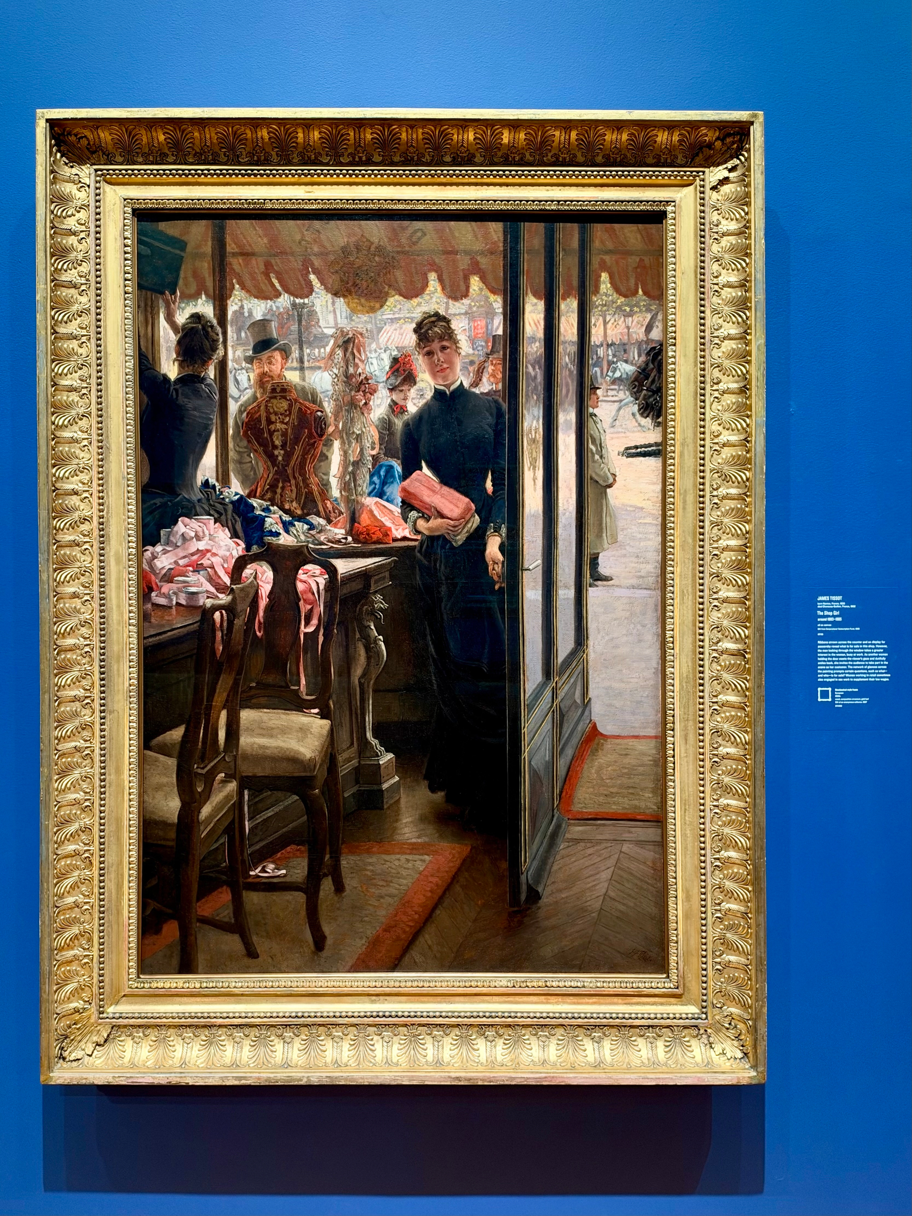A framed painting hangs on a vibrant blue wall in a gallery. The painting depicts the interior of a shop with a woman in a dark dress holding folded pink fabric, standing in the doorway. Behind her, through the shop window, several figures in period clothing can be seen, including a man in a decorated coat and hat and other women wearing elaborate hats. Inside the shop, a wooden chair with a cushioned seat faces a table stacked with various colourful fabrics, primarily pinks and blues. The shop has wooden floors with patterned rugs, and light from the outside brightens the scene. The ornate golden frame features intricate carved details, including a repeating leaf motif. To the right of the painting, a small wall plaque with white text provides information about the artwork, though it is not legible in the image.