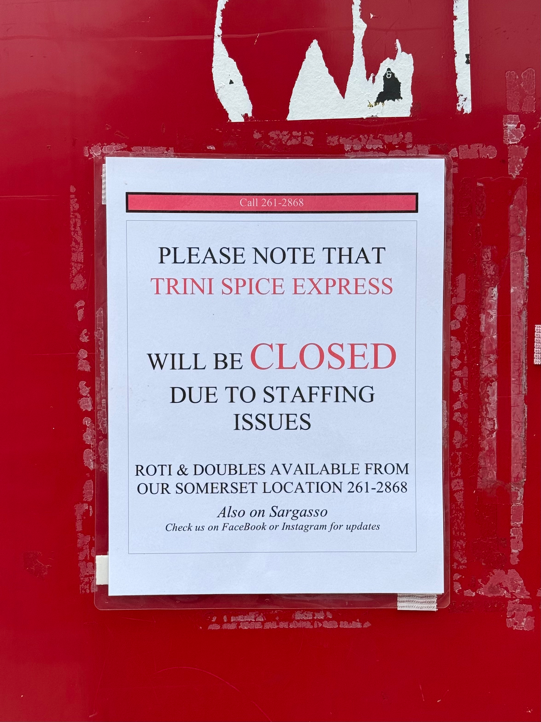 A printed notice is taped to a bright red, weathered surface with remnants of old tape and peeling white stickers. The notice has a white background with a red header and black and red text. The header reads “Call 261-2868.” The main body of the notice says, “PLEASE NOTE THAT TRINI SPICE EXPRESS WILL BE CLOSED DUE TO STAFFING ISSUES.” The word “CLOSED” is in bold red letters, with the rest of the text in black. Below, it states, “ROTI & DOUBLES AVAILABLE FROM OUR SOMERSET LOCATION 261-2868.” At the bottom, it says in italics, “Also on Sargasso” and “Check us on FaceBook or Instagram for updates.” The edges of the sign are clear-taped to the red surface, and the lower right corner of the paper is slightly curled.