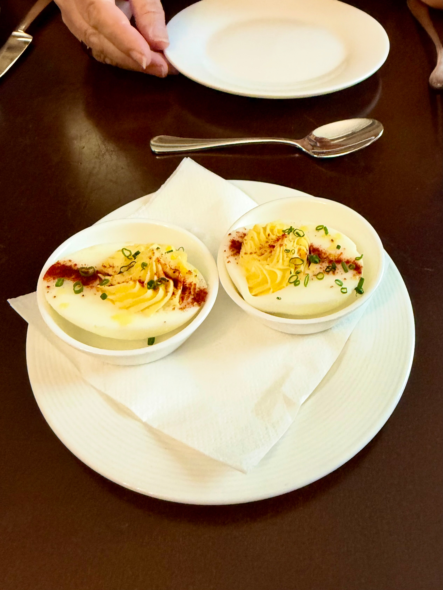 Two devilled eggs served on small white dishes, placed on a white plate with a folded napkin. The eggs are garnished with chopped green herbs and a sprinkling of red paprika. In the background, there is a silver spoon, a white plate, and a person's hands resting on the table. The table is dark brown, providing contrast to the white dishes.